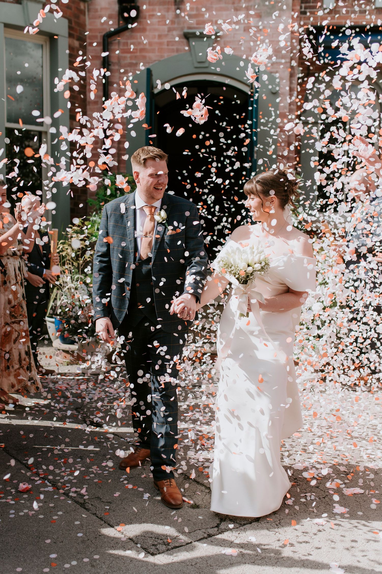 Wedding couple celebrates under a shower of pink and white confetti outside a brick building.