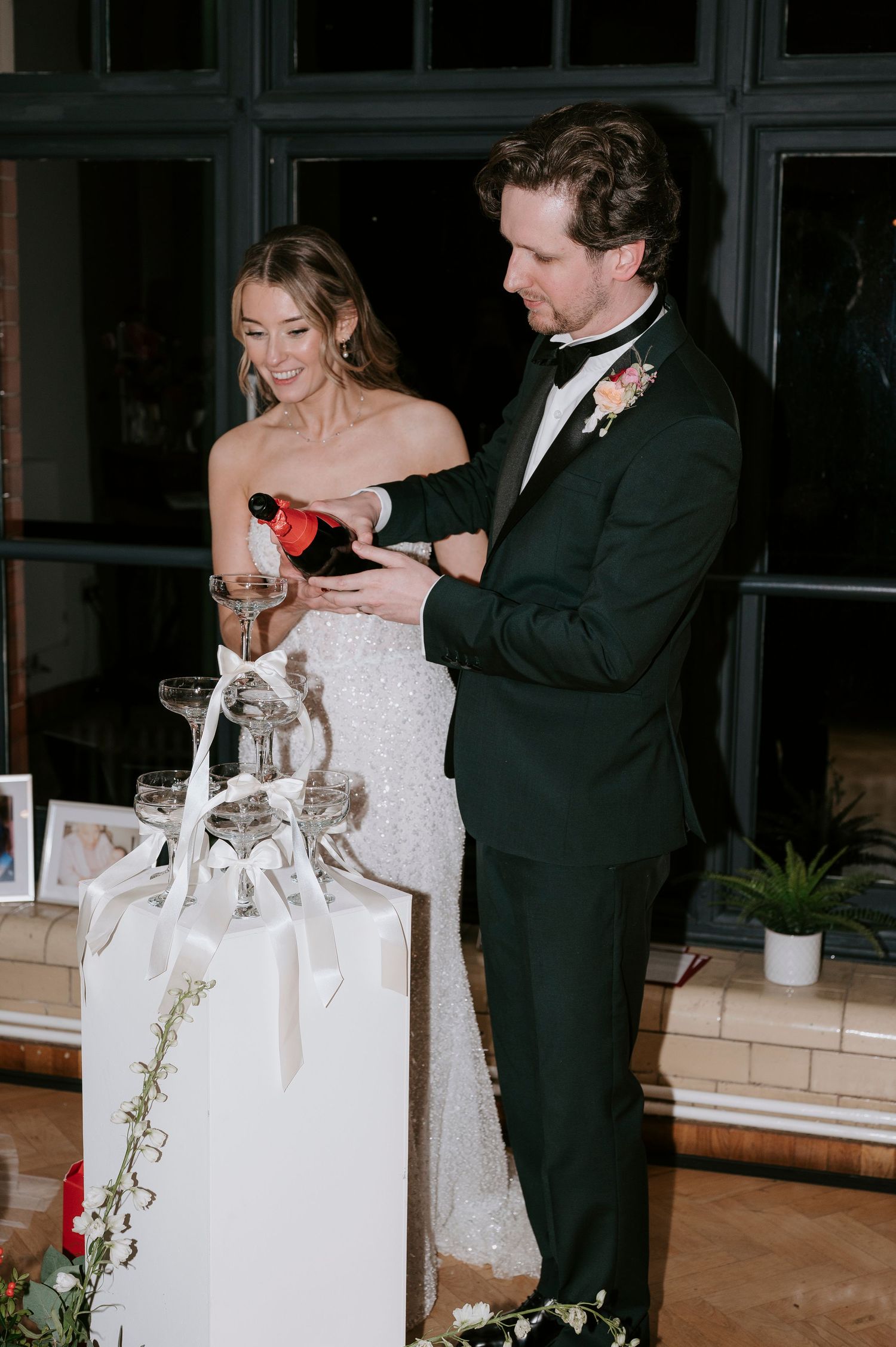 A bride in a white gown and groom in a tuxedo pour champagne together during their wedding reception.