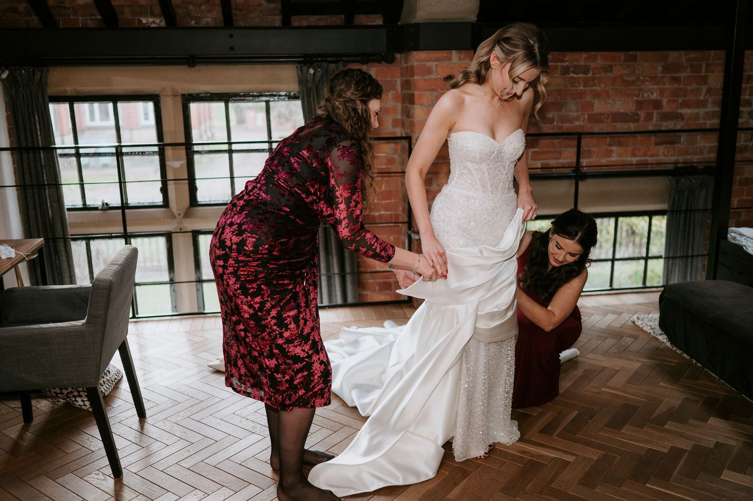 A bride in a white wedding dress stands in a rustic brick-walled room with warm lighting and hardwood floors.