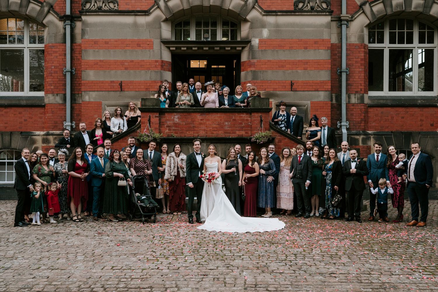 Wedding guests line up outside red brick building for group photo on cobblestone street.