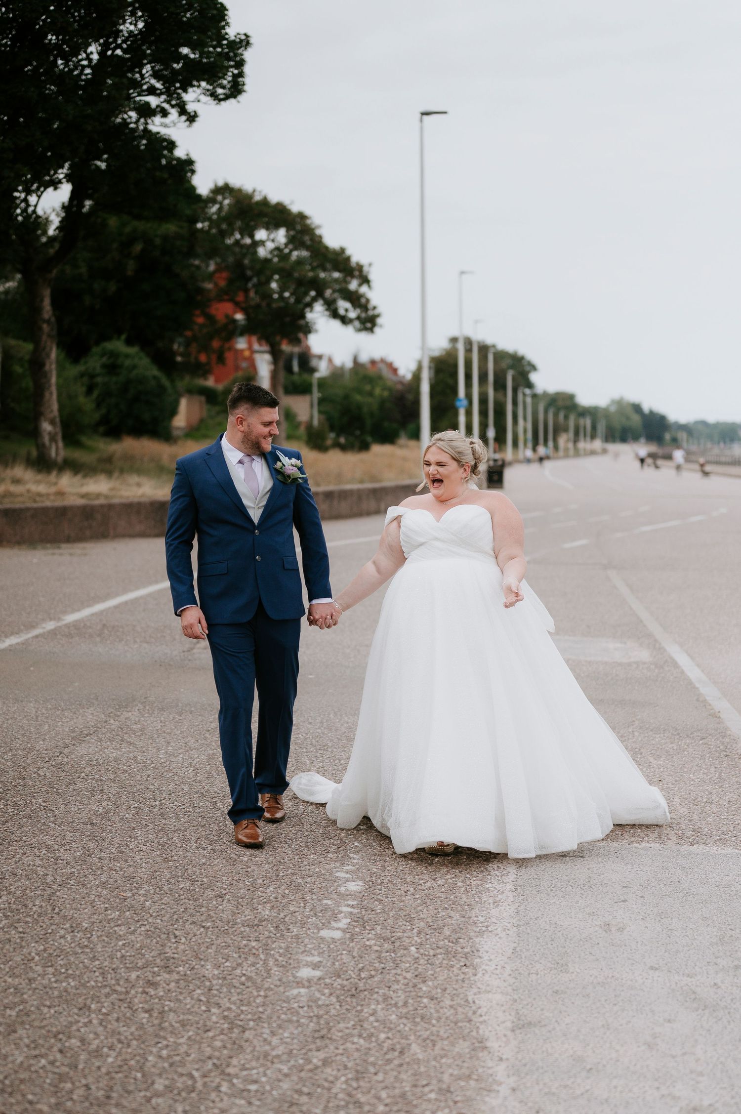 A couple in wedding attire walk and dance together on a paved road lined with trees and lampposts.