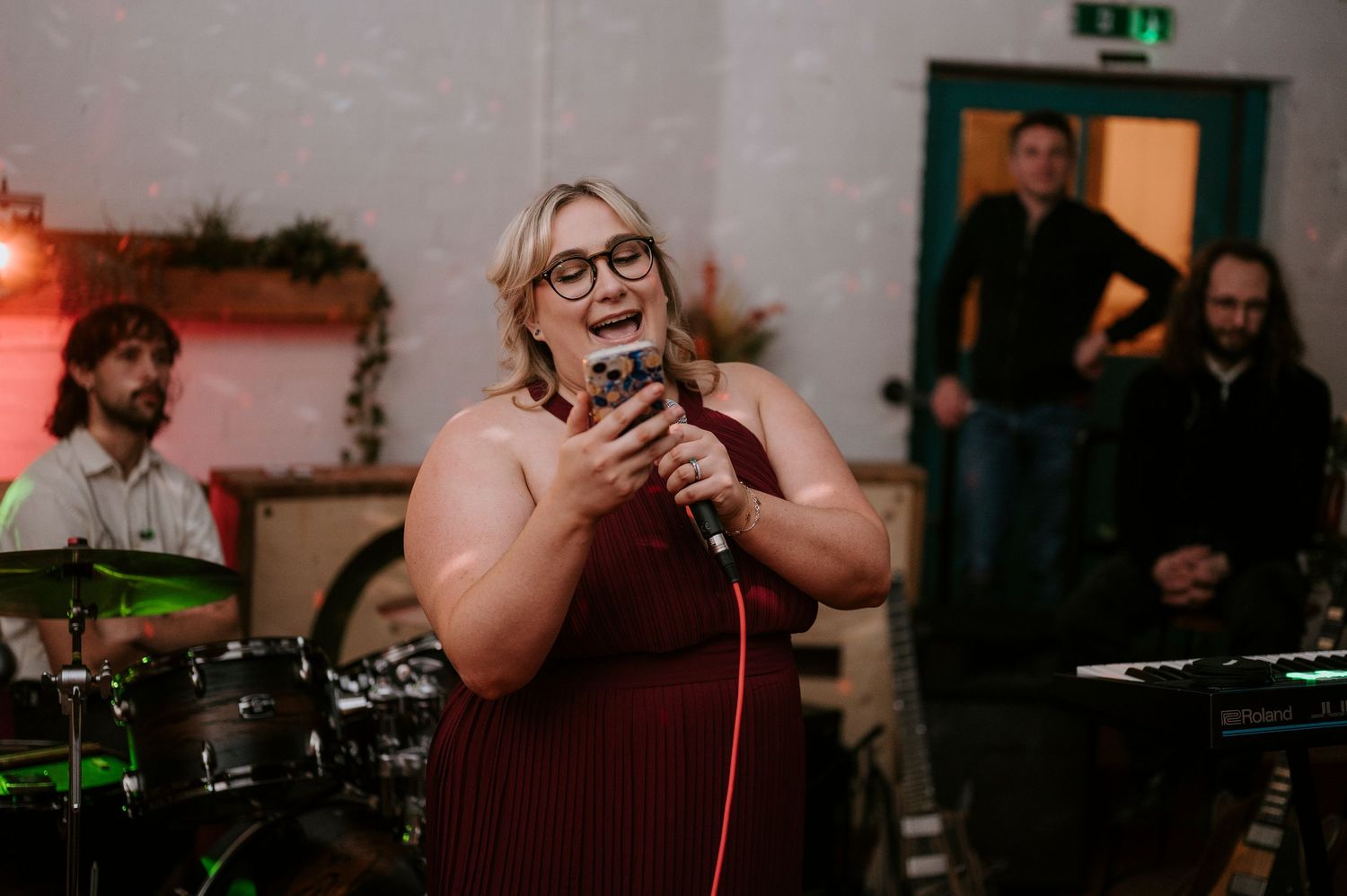 A woman with glasses singing into a microphone at an indoor event with band members visible in the background.