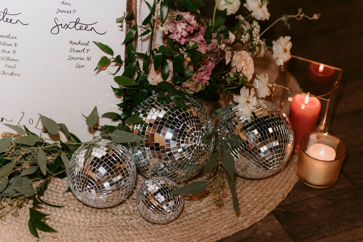 Disco balls and lit candles arranged with pink flowers and greenery on a natural woven mat create an elegant party setting.