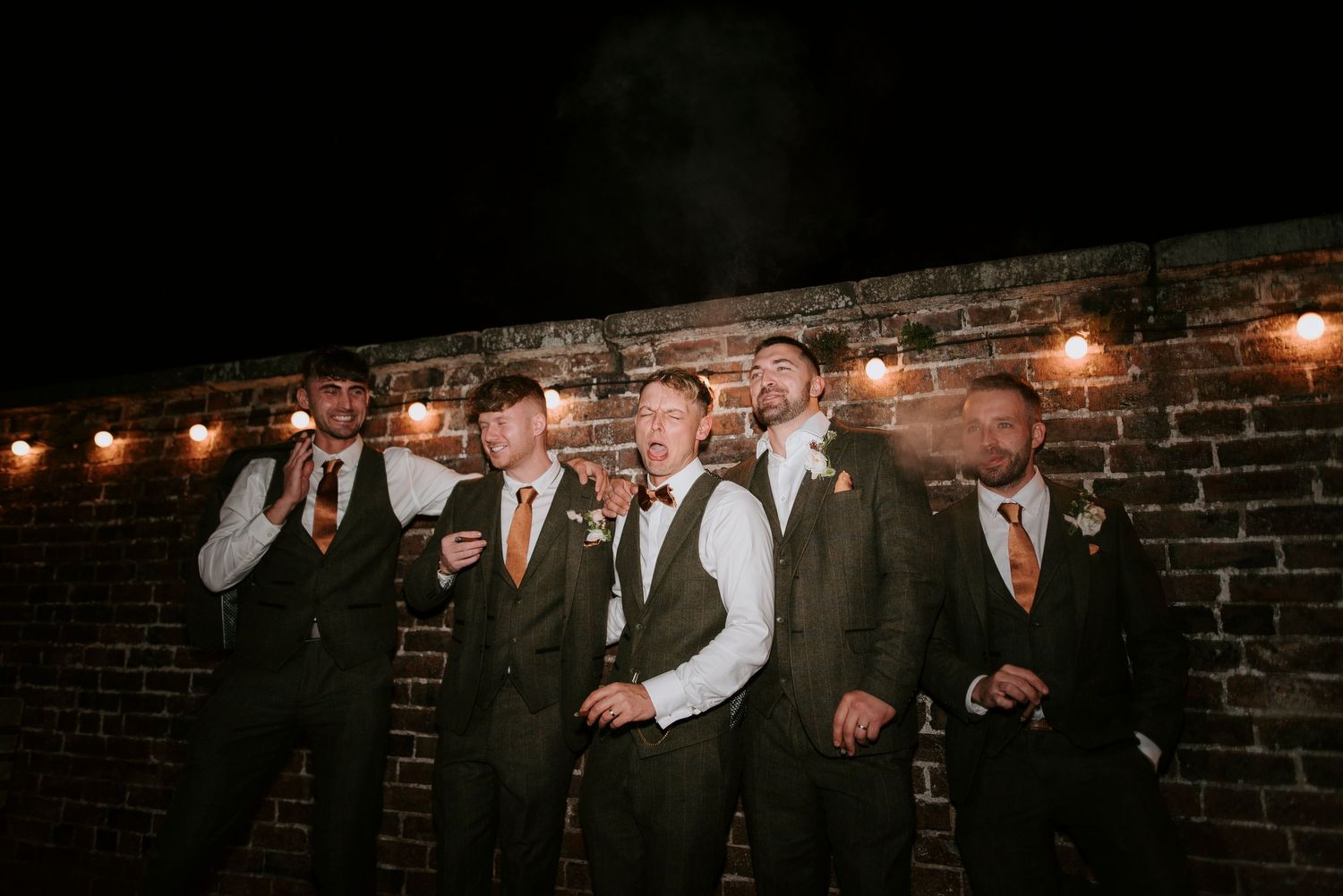 A group of people in formal attire stand against a rustic brick wall with string lights overhead at an event.