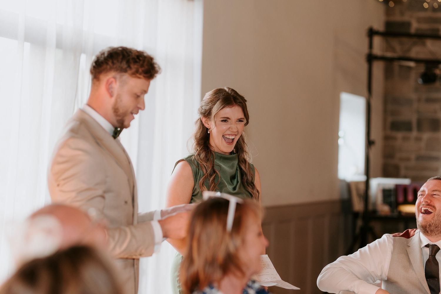 Wedding guests laugh and celebrate during a joyful reception in a rustic indoor venue with brick walls.