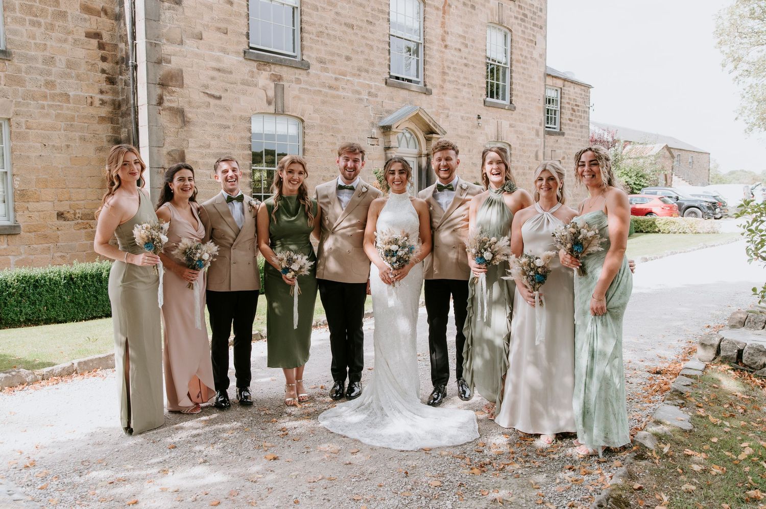 Wedding party wearing neutral tones and sage green dresses poses together outside stone building.