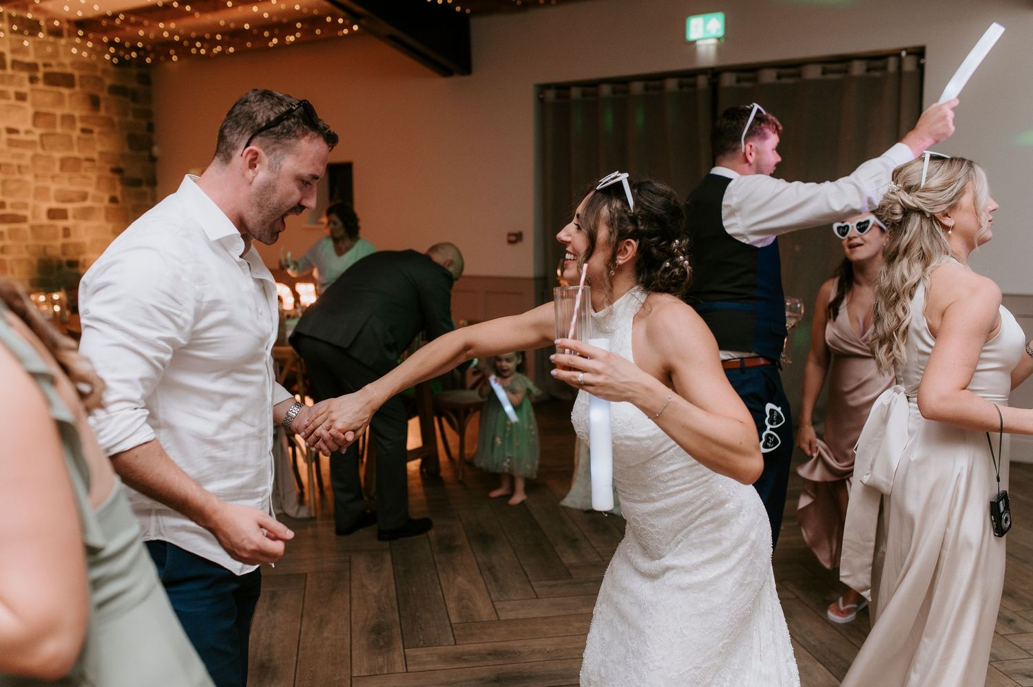 Wedding guests dance energetically in a rustic venue decorated with warm string lights and exposed brick walls.