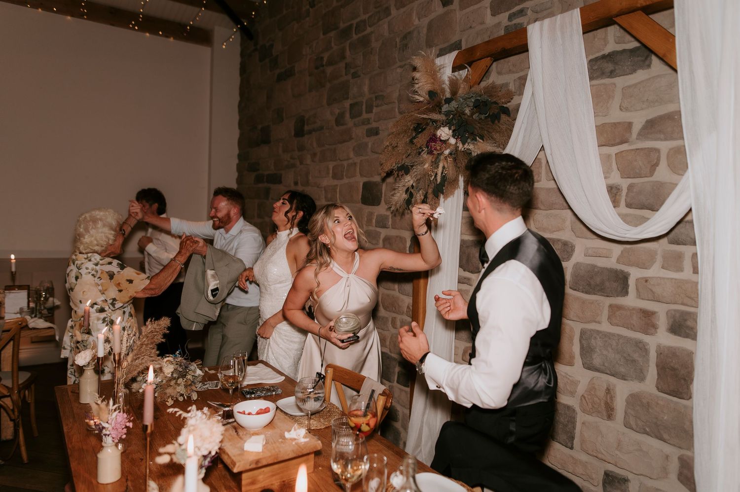 Wedding reception guests dance and celebrate together on a dance floor in a rustic brick-walled venue.