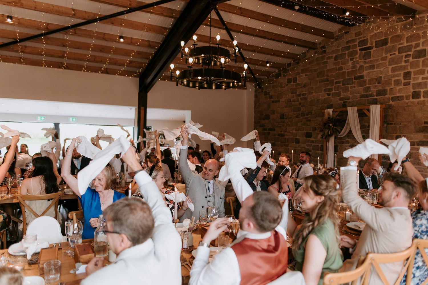 Wedding reception guests wave white napkins in celebration during a joyful moment at a rustic brick venue.