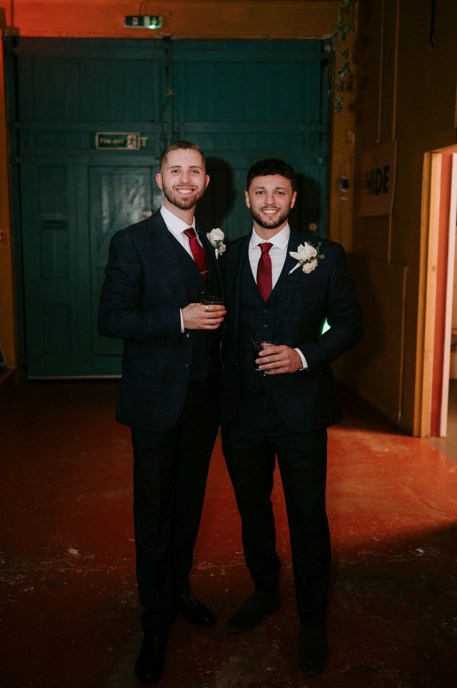 Two grooms in navy suits with red ties and boutonnieres pose together at their wedding reception.