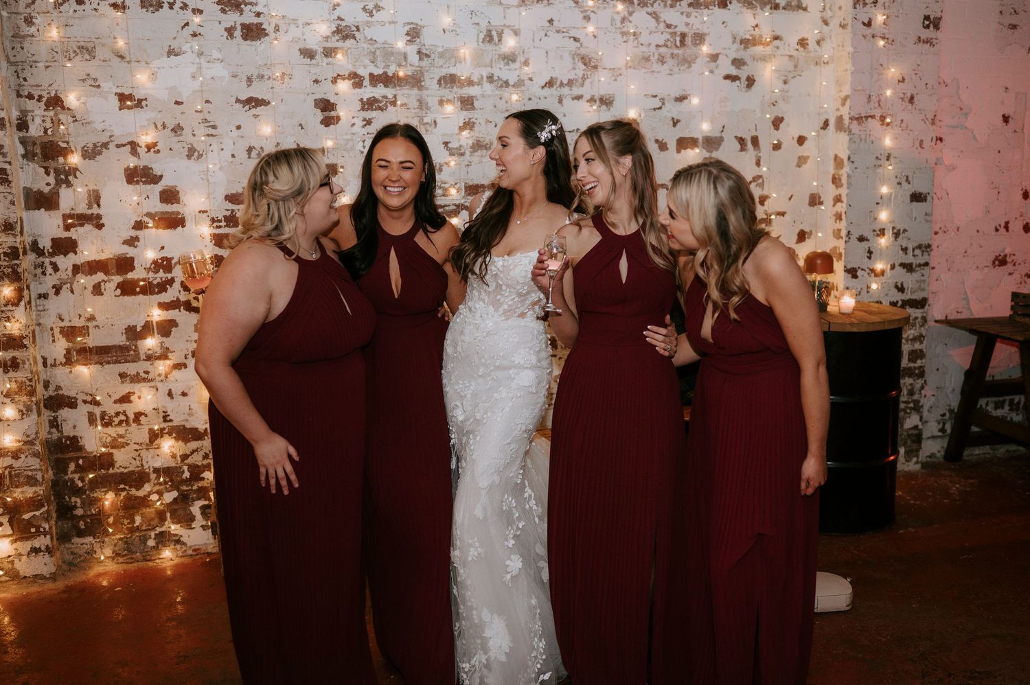 Bride in white lace gown poses with four bridesmaids in burgundy dresses against a twinkling light backdrop.