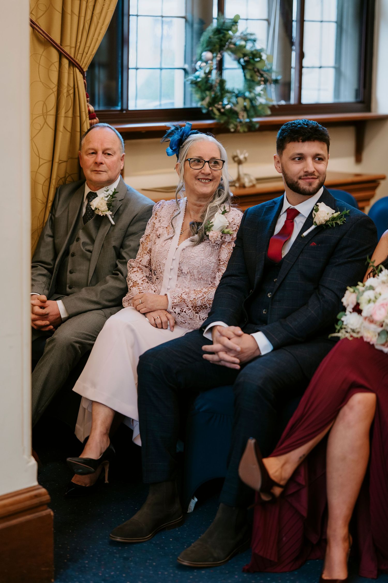 Wedding guests smiling during ceremony, woman in blue fascinator hat seated between two men in suits.