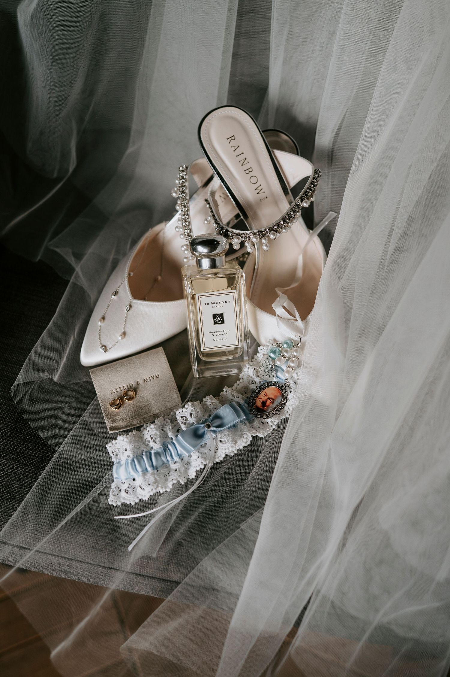 Elegant bridal accessories including white heels, perfume bottle, and jewelry arranged on white tulle fabric.