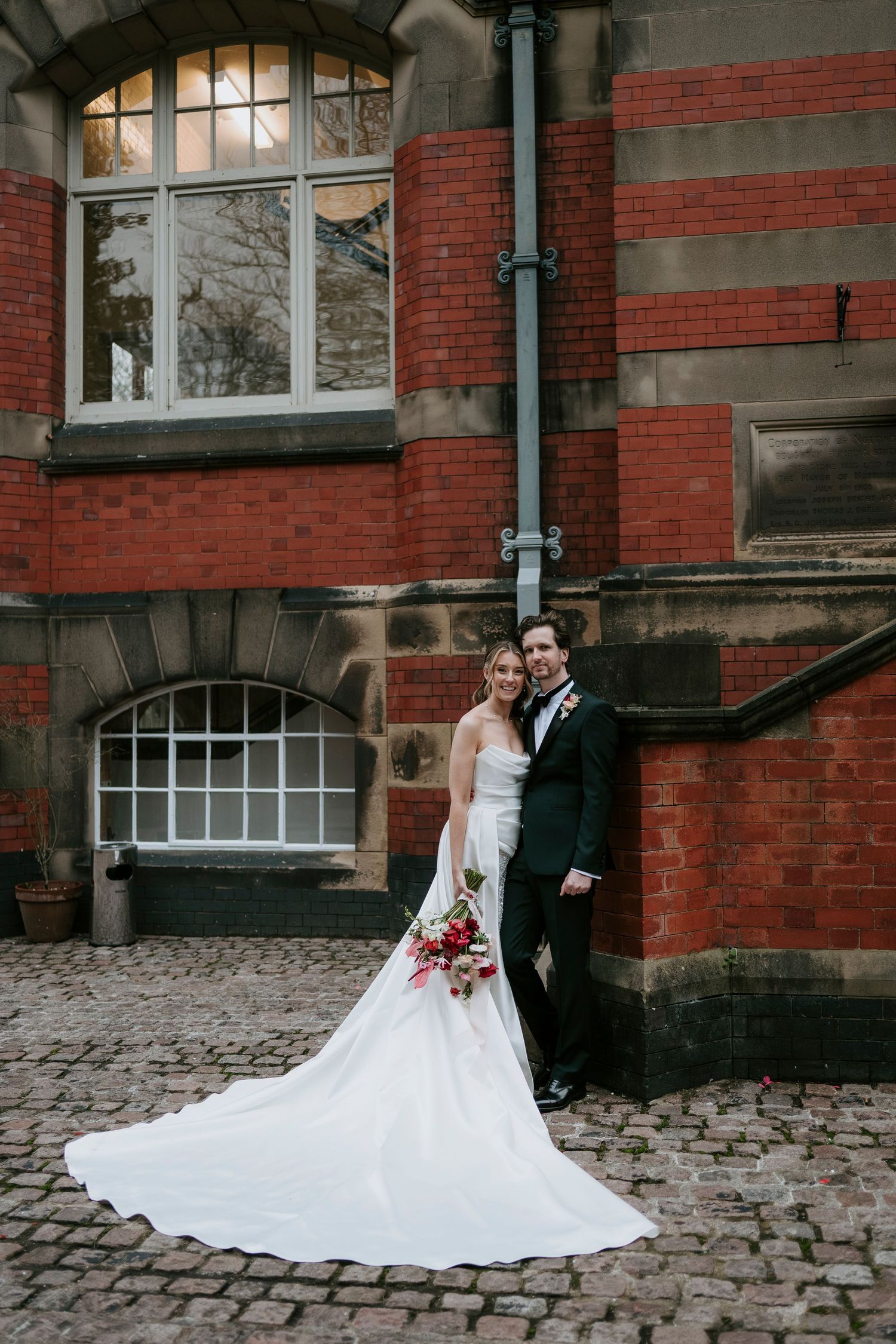 Elegant wedding couple poses on cobblestone path against historic red brick building.
