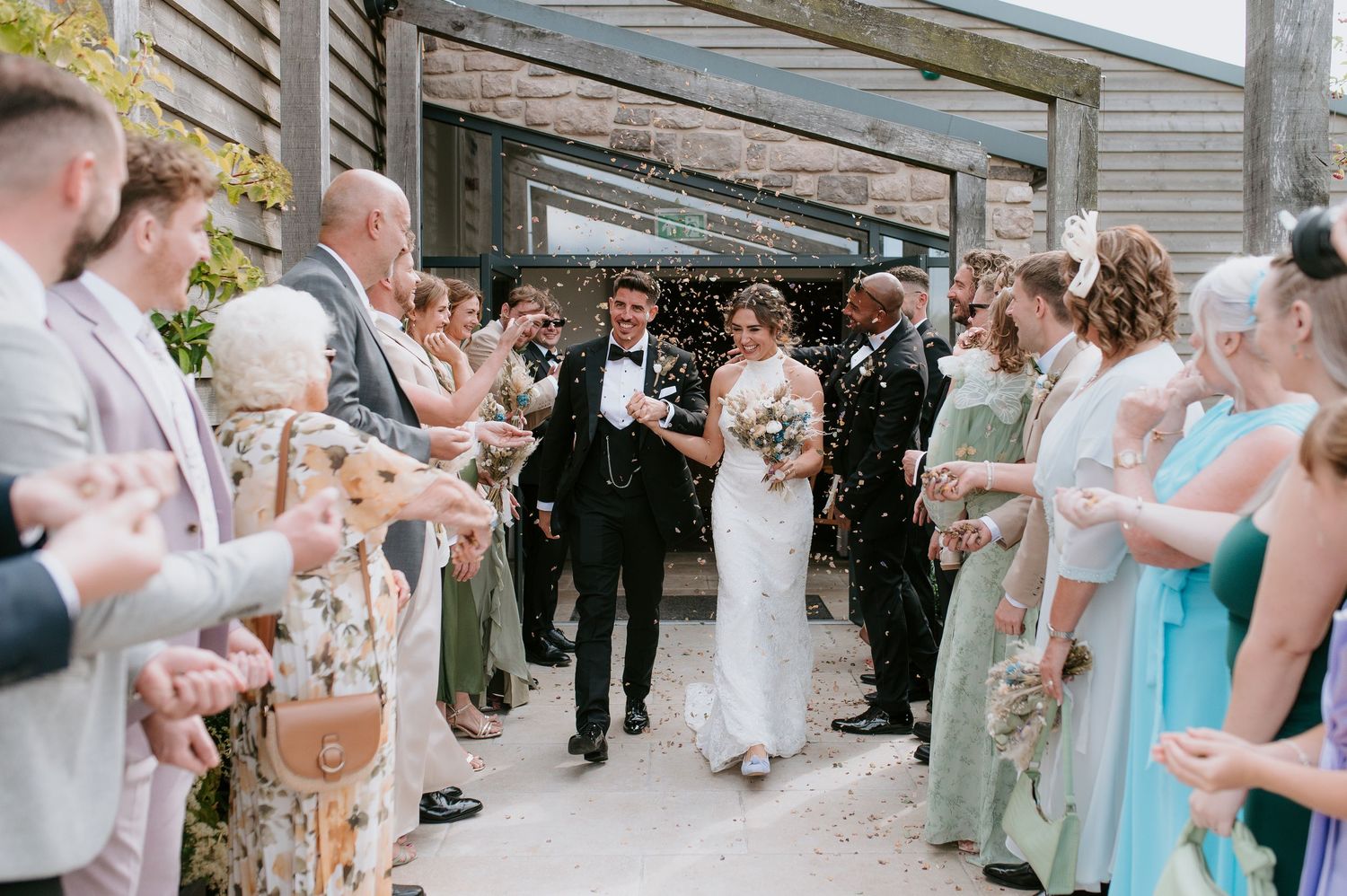 Wedding guests line a walkway blowing bubbles as a couple exits through an outdoor venue path.