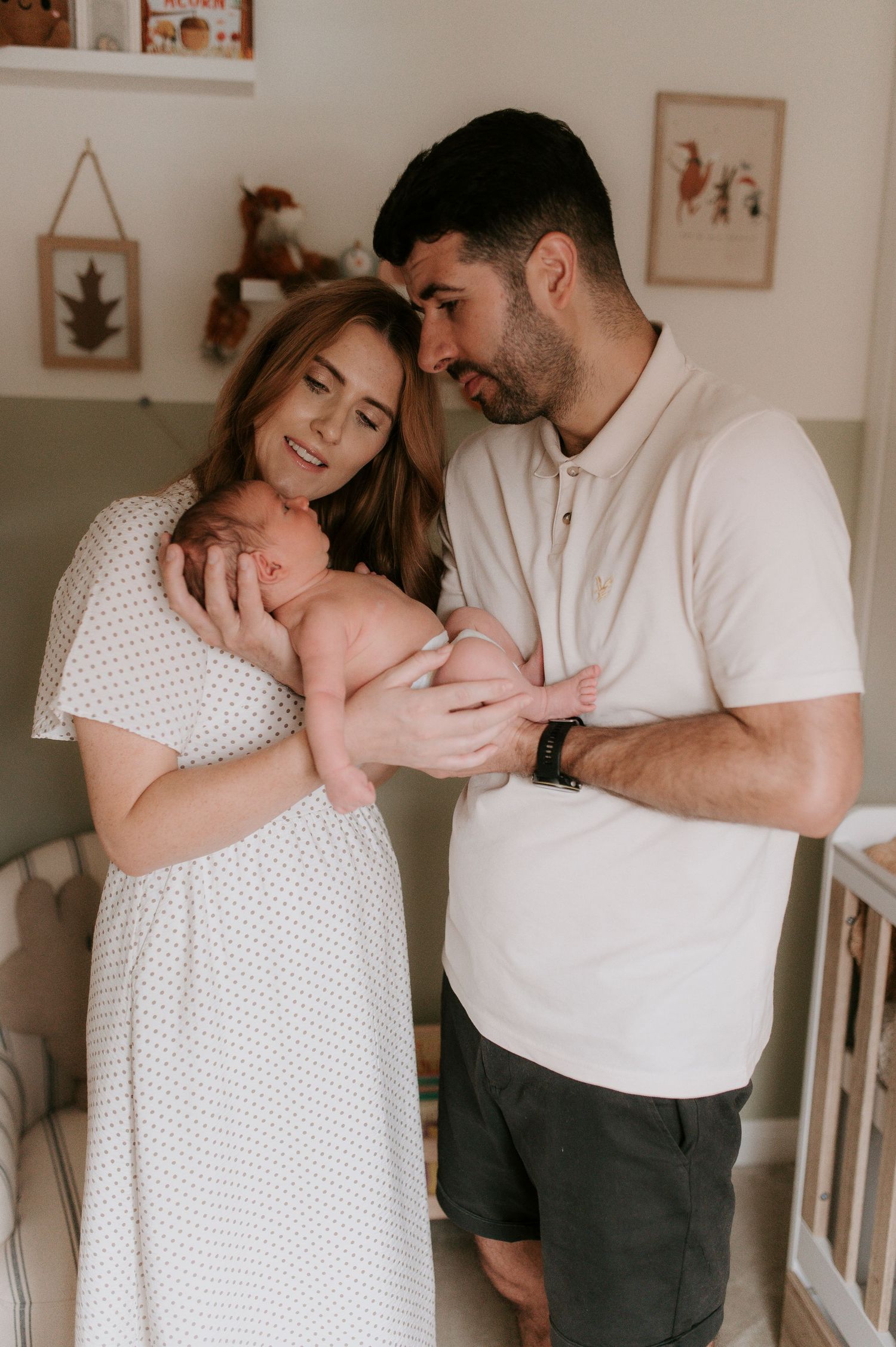 A couple in white clothing tenderly holds their newborn baby in a nursery setting with wall art visible in background.