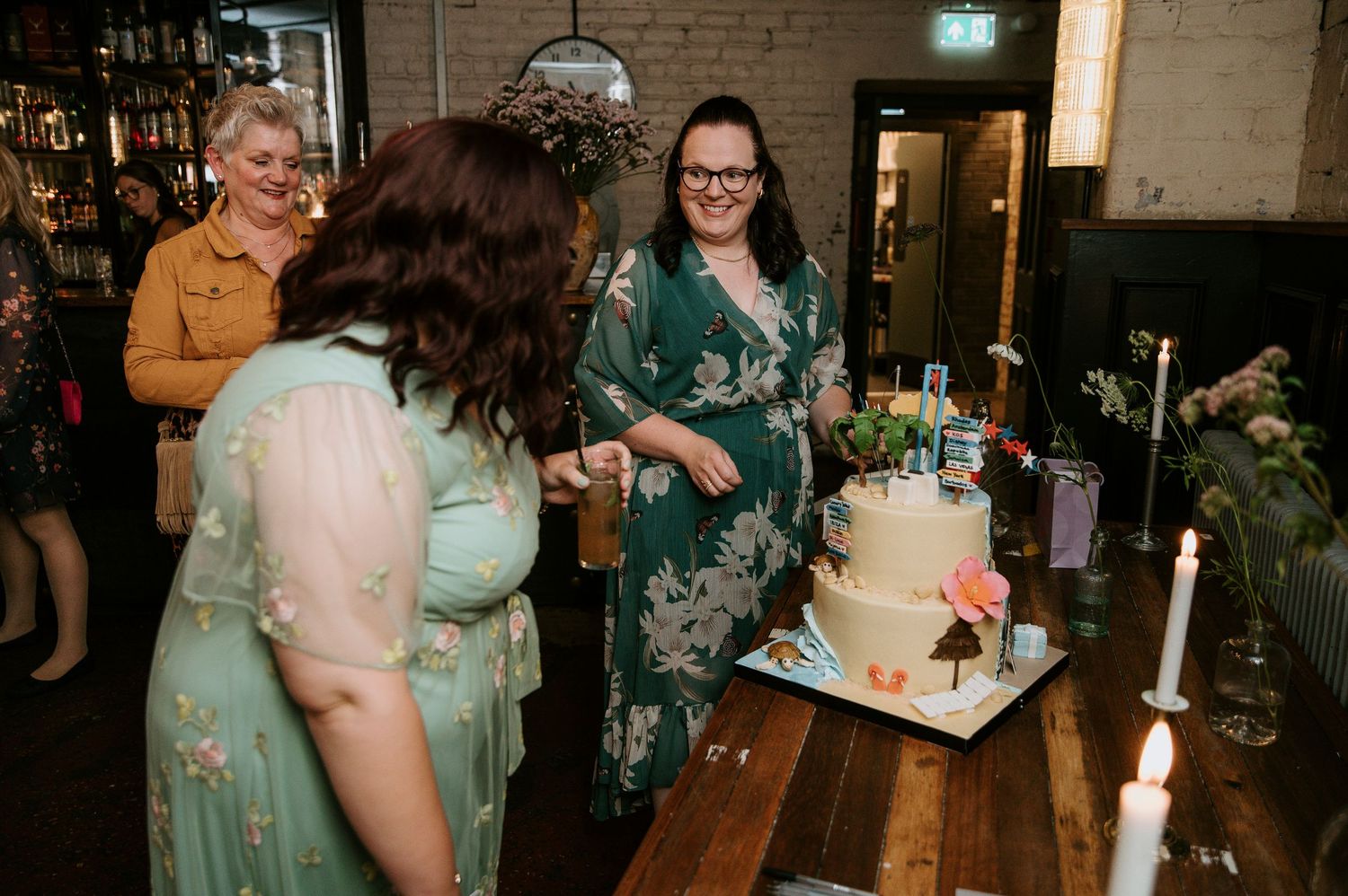 A group gathers around an elegant white wedding cake adorned with flowers and candles on a wooden table at a reception.