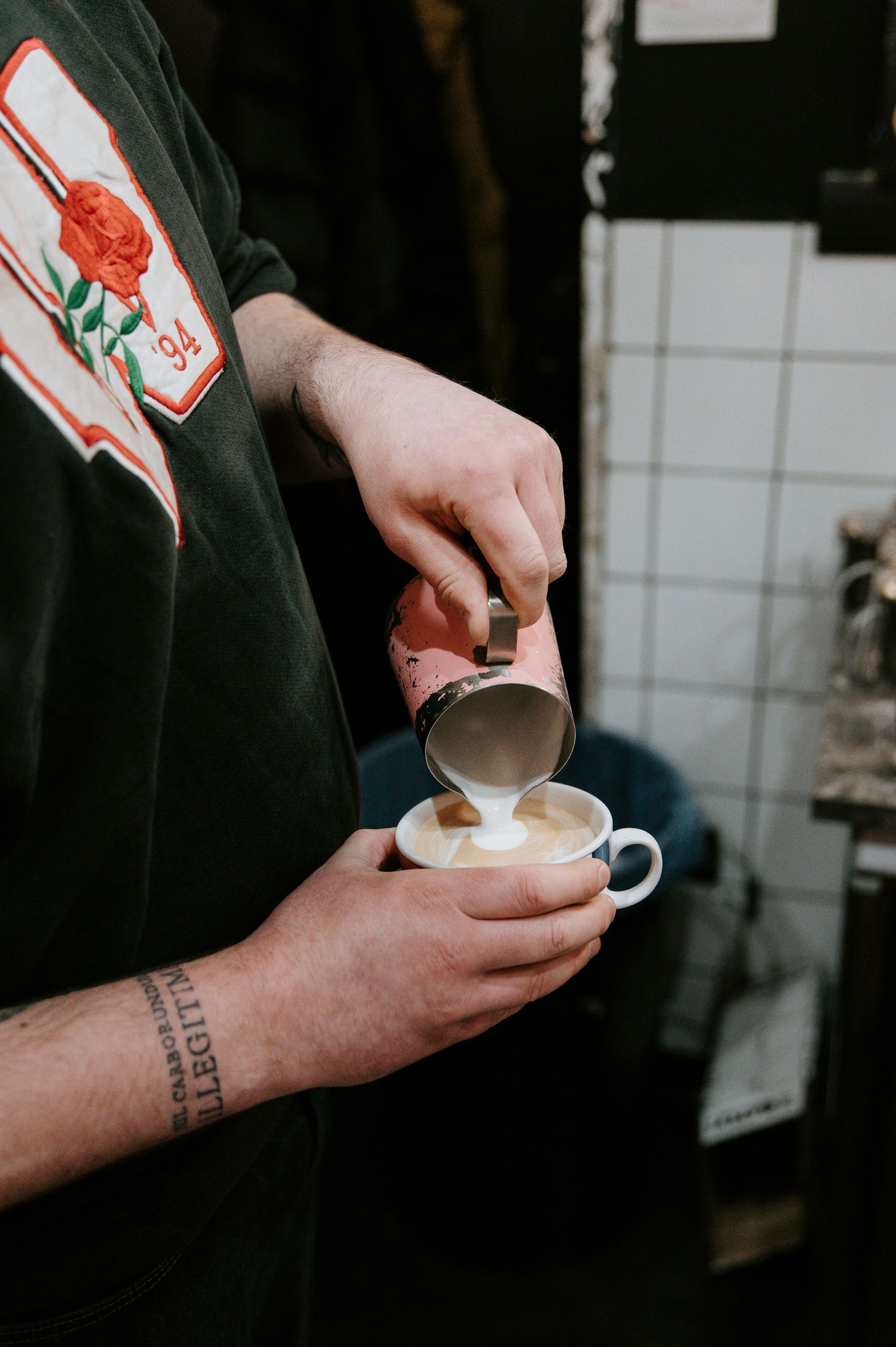 A tattooed hand pours steamed milk into a small white coffee cup while wearing a black shirt with a red logo patch.