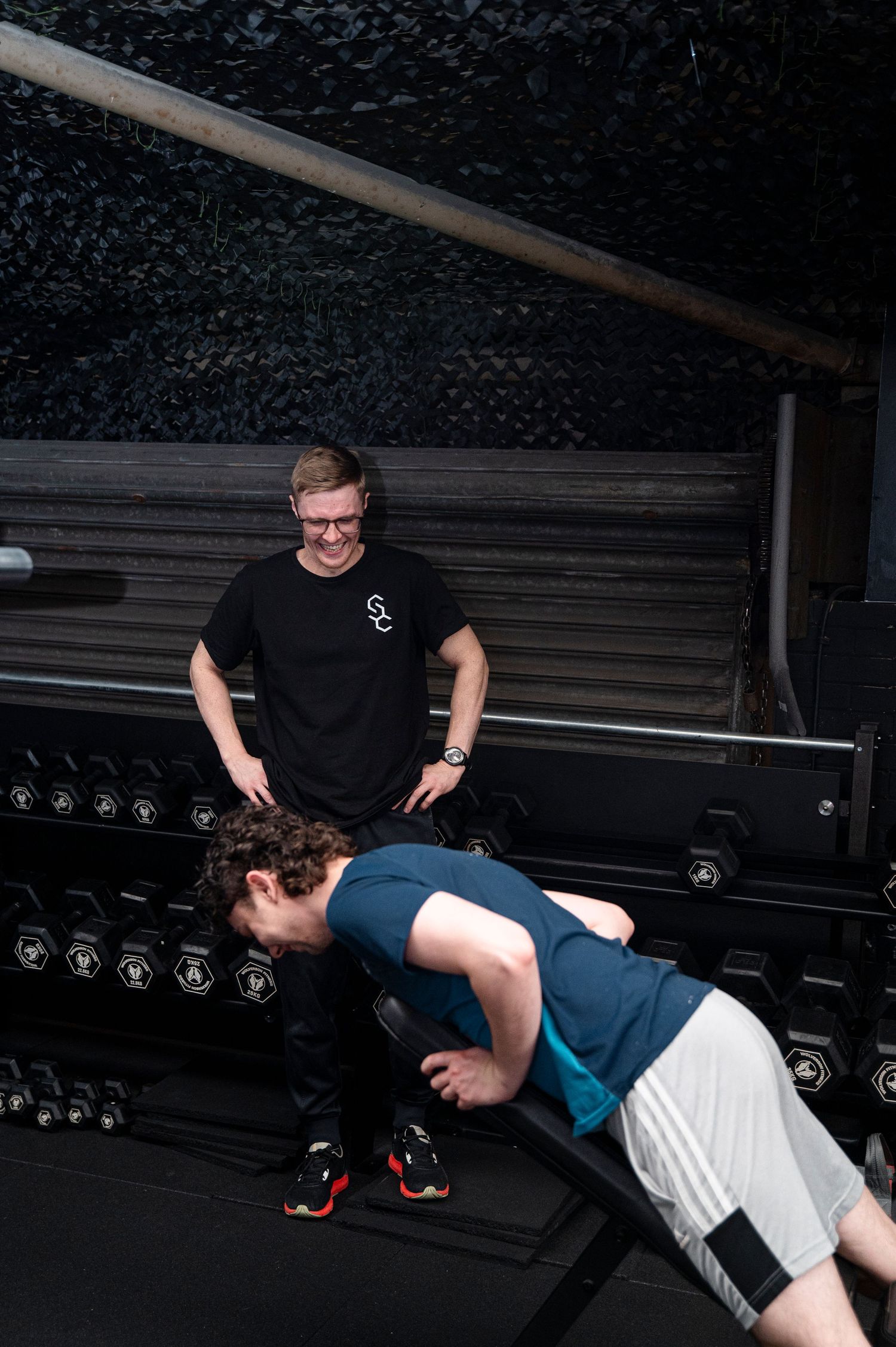 A fitness instructor guides someone during a bench press exercise in a dimly lit gym facility.