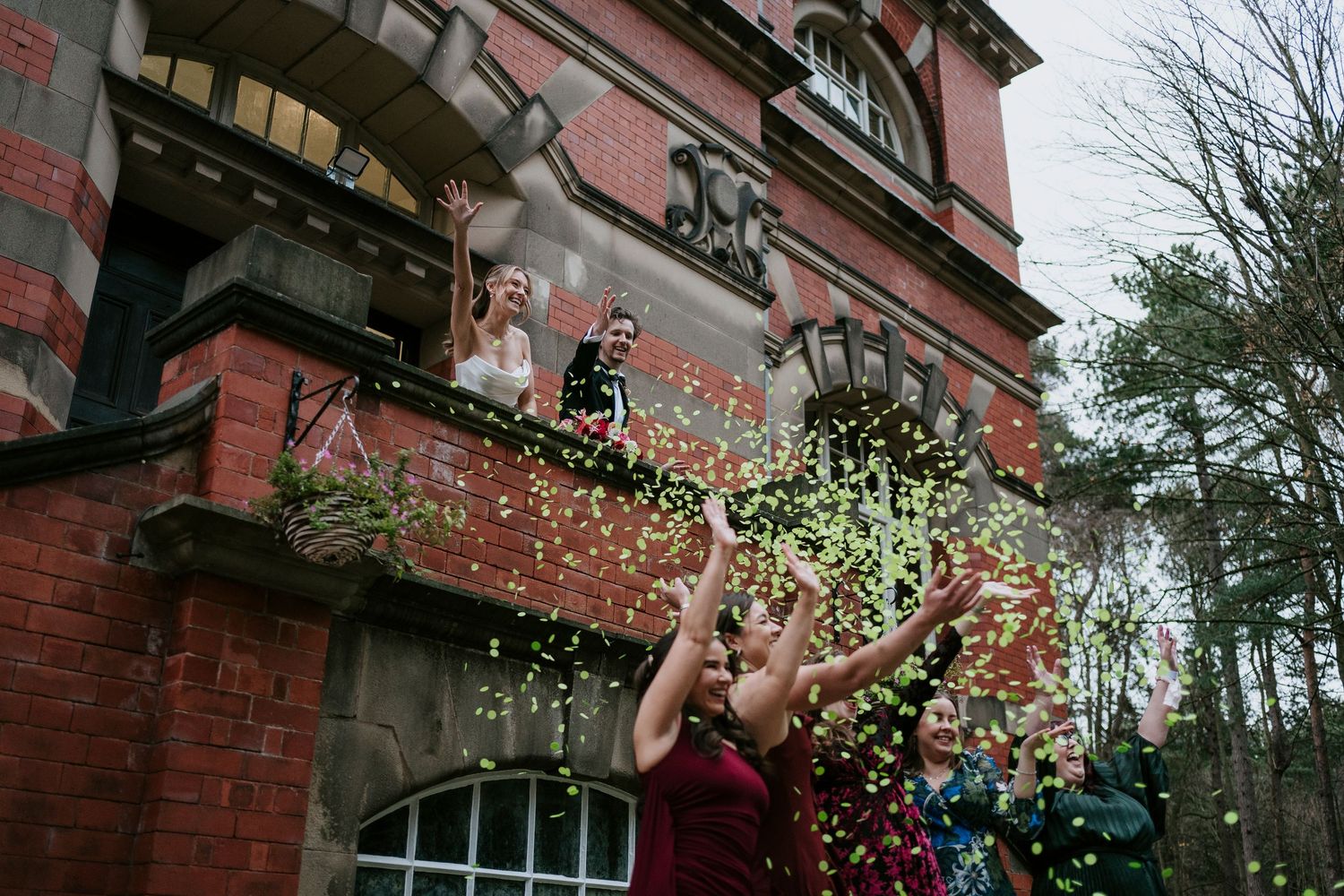Colorful confetti rains down against red brick architecture during outdoor wedding celebration.