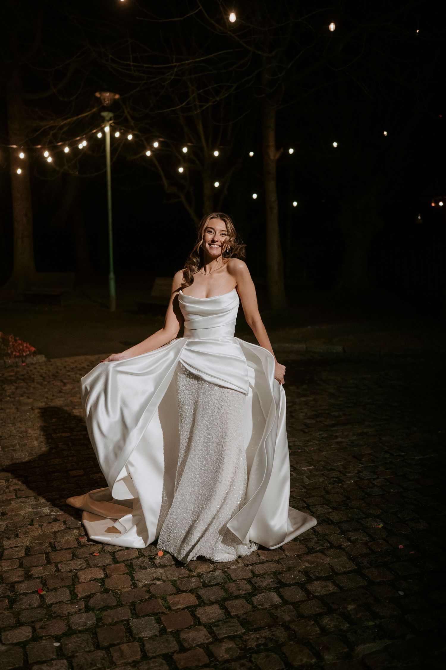 A bride in a stunning white gown poses elegantly on a cobblestone path under string lights at night.