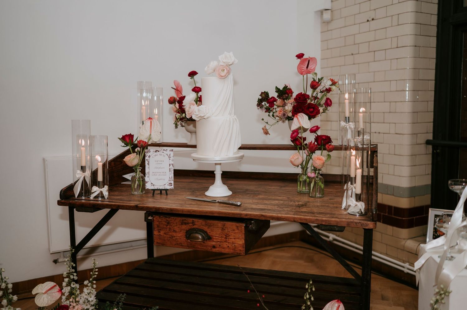 A rustic wedding reception setup with wooden tables, white vases, and red floral arrangements against brick walls.