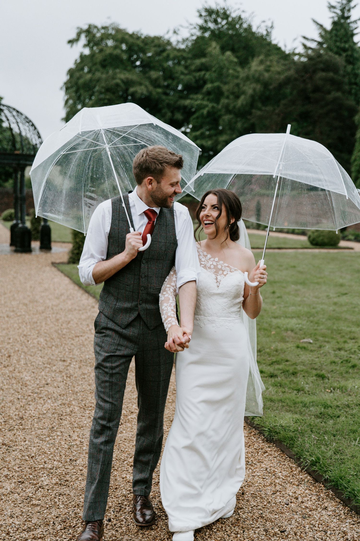A couple shares a joyful moment under clear umbrellas on a rainy wedding day along a gravel path.