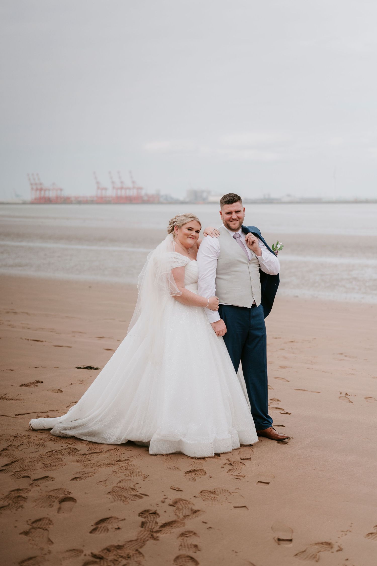 A wedding couple poses together on a sandy beach with industrial cranes visible in the misty background.
