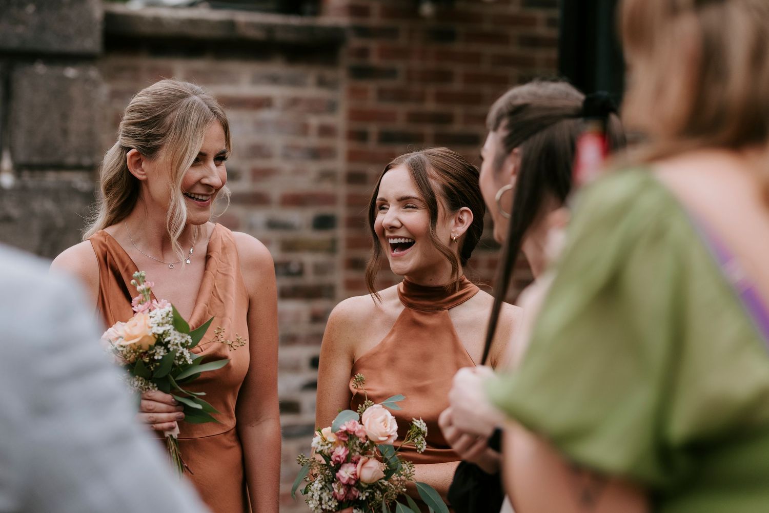 Joyful wedding party sharing candid laughter in front of a brick wall during golden hour.
