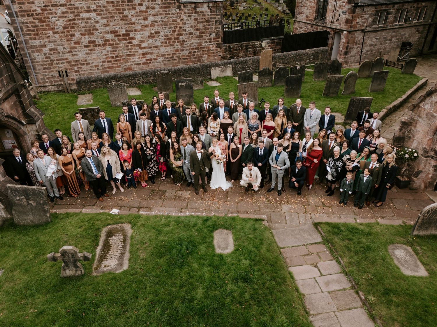 Large wedding party group photo taken at a rustic brick church venue with stone pathways and green lawns.