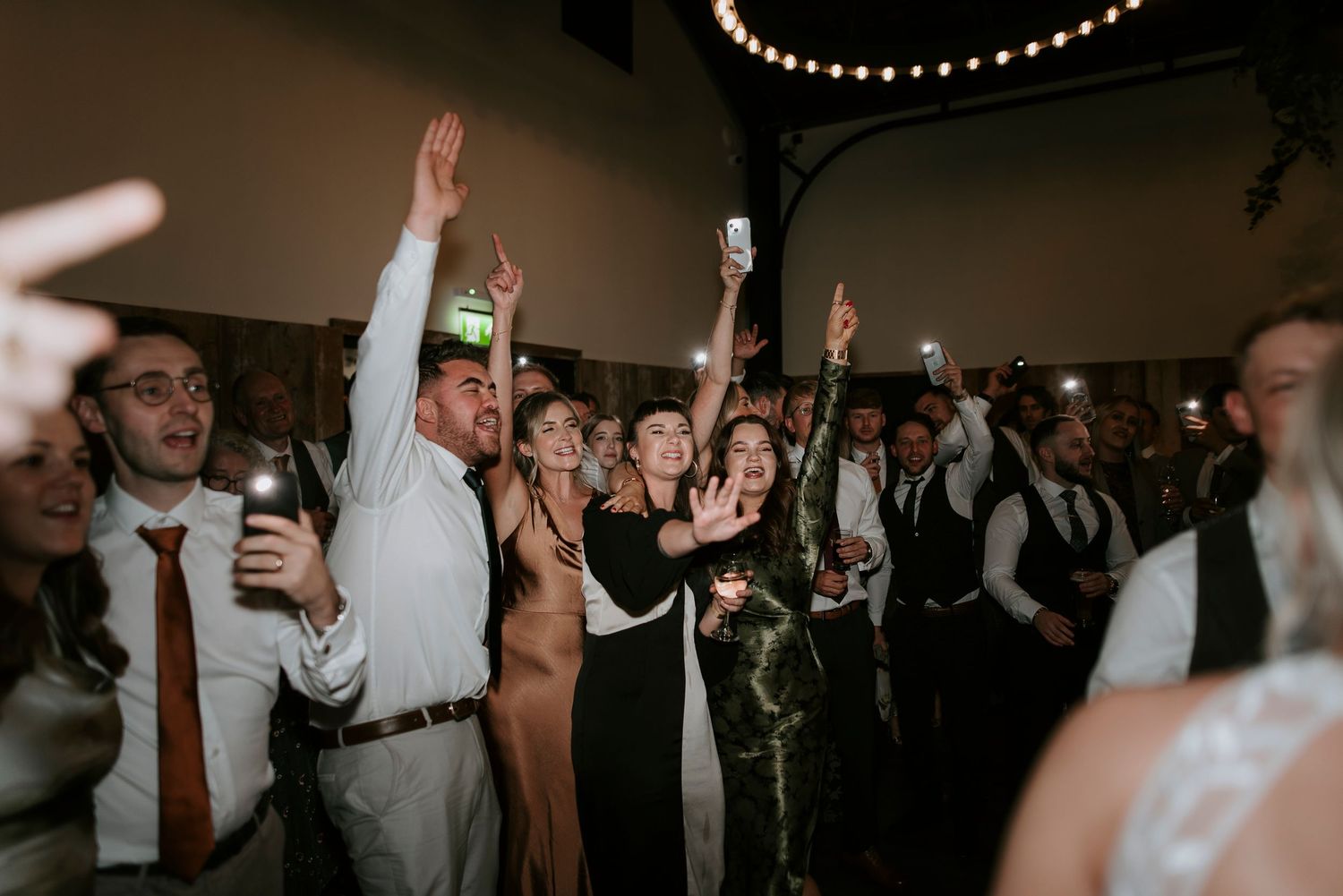 Wedding reception guests dancing and celebrating with raised hands on a dark dance floor at an indoor venue.