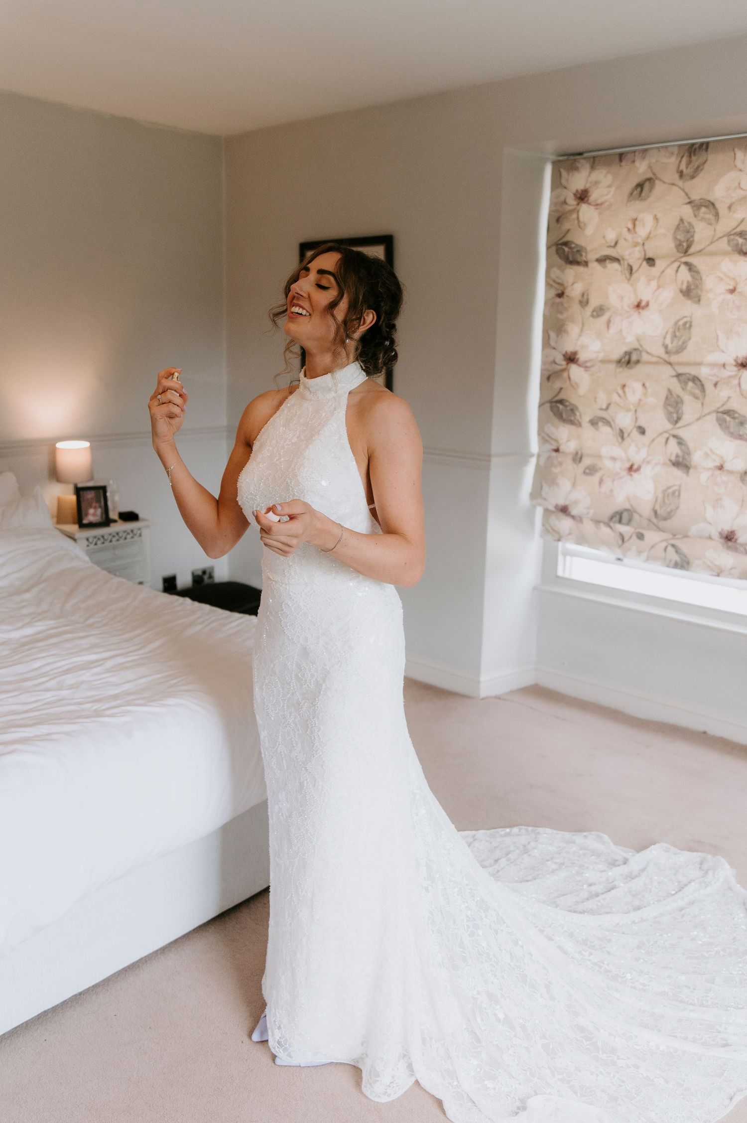 A bride in a flowing white lace wedding dress poses playfully on a bed in a light-filled bedroom.