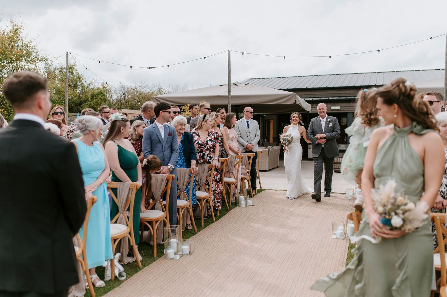 Outdoor wedding ceremony with guests seated on wooden chairs along a paved aisle under string lights.