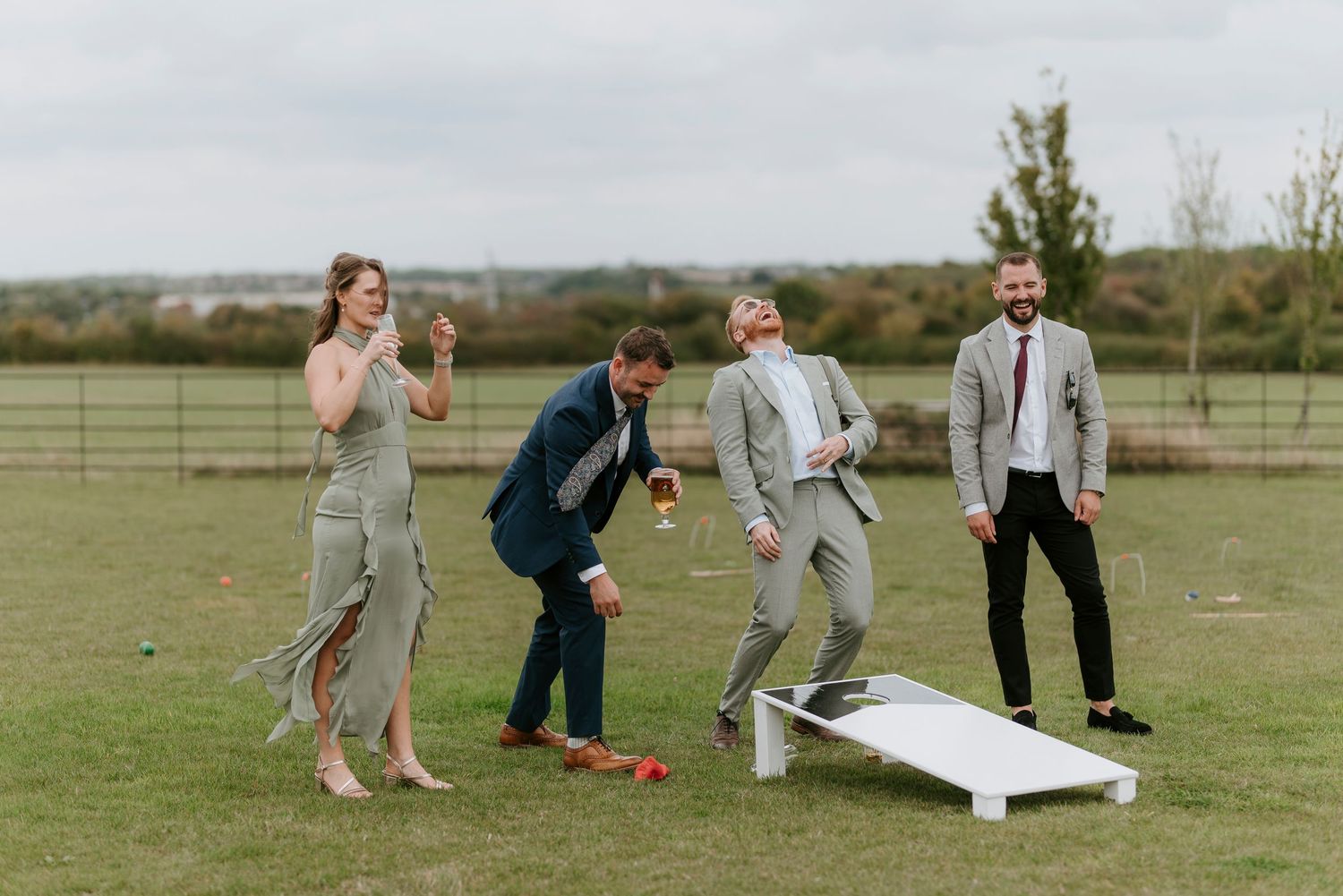 People playing cornhole lawn game outdoors at a casual event wearing formal attire.