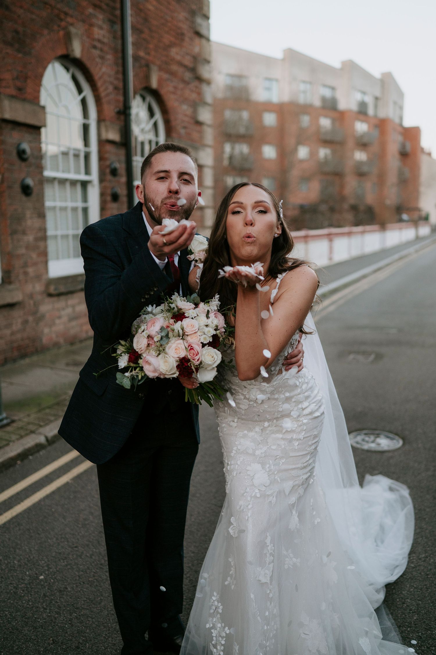 Bride and groom blowing confetti on a street, bride holding a pink bouquet in a lace gown with veil.