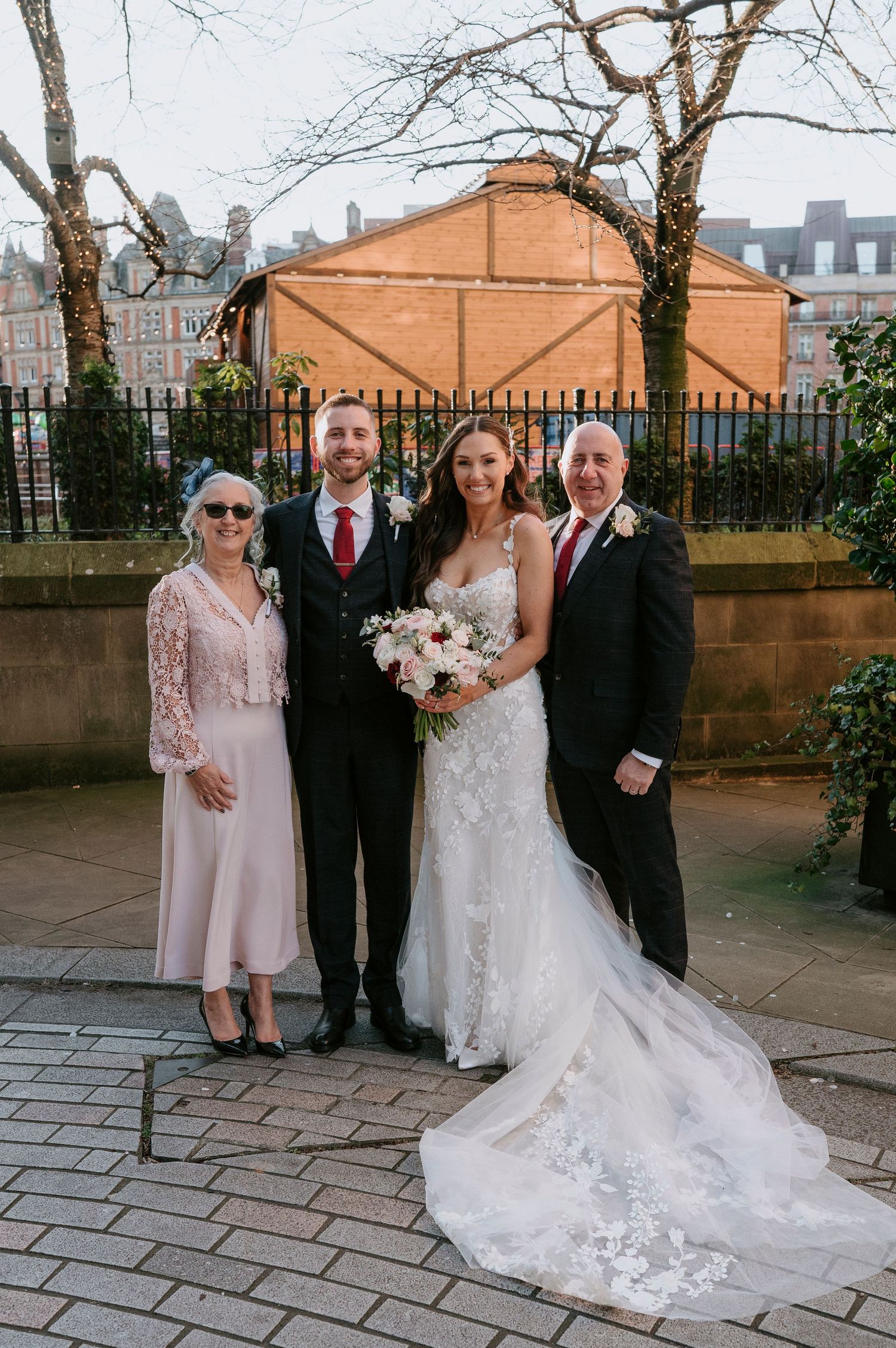Bride and groom pose with two family members outdoors, bride holding bouquet in lace mermaid gown.
