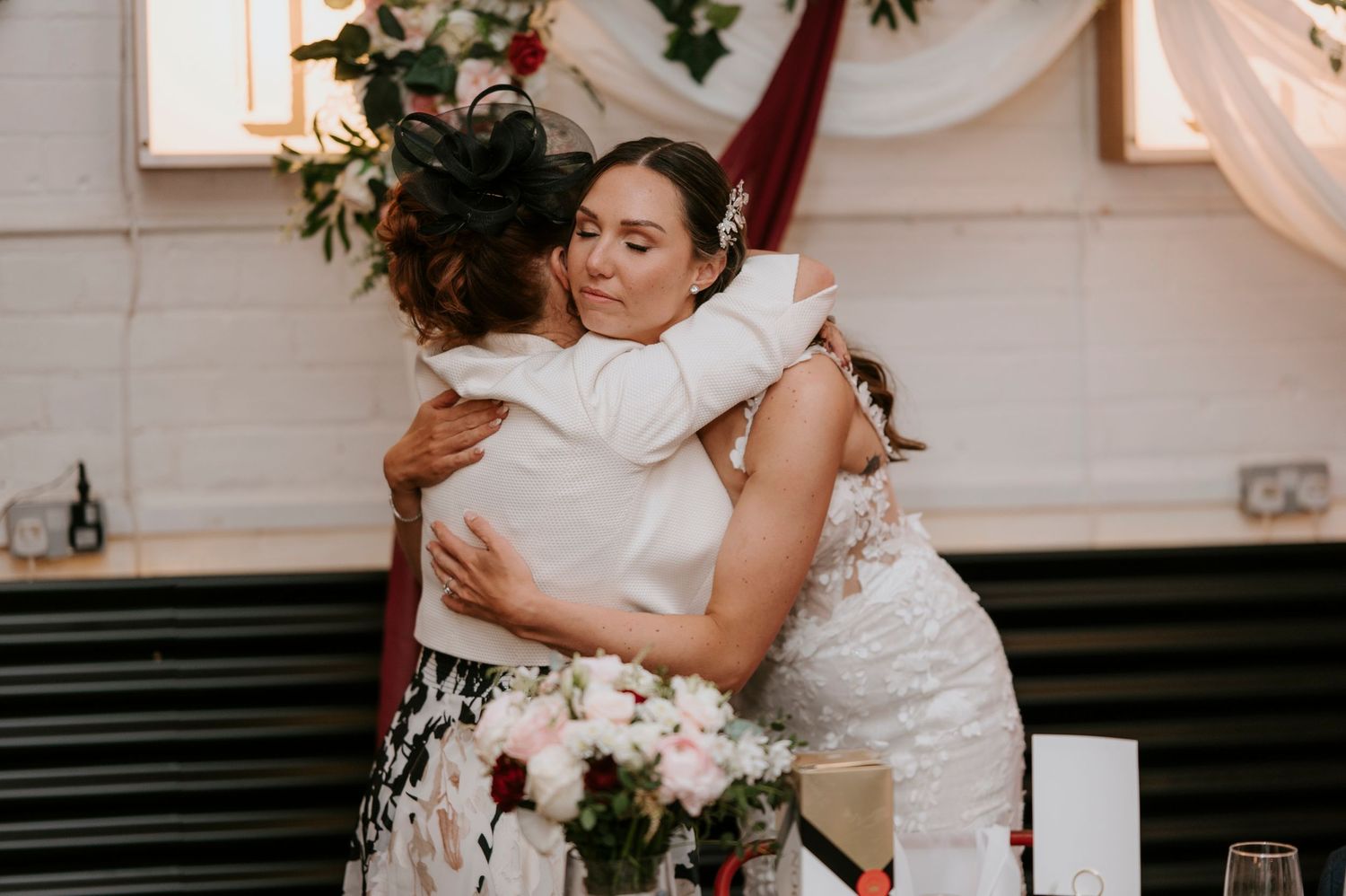 Bride shares emotional hug with guest in floral dress at wedding reception with floral centerpieces.