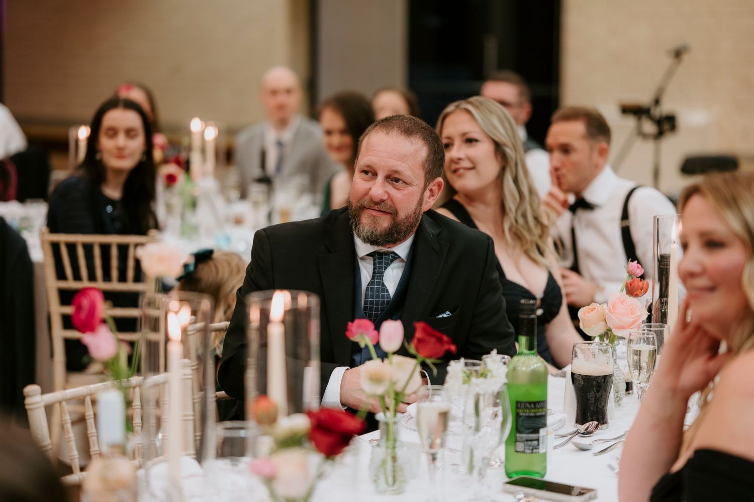Wedding guests enjoy conversation and drinks at an elegant reception dinner table decorated with floral centerpieces.