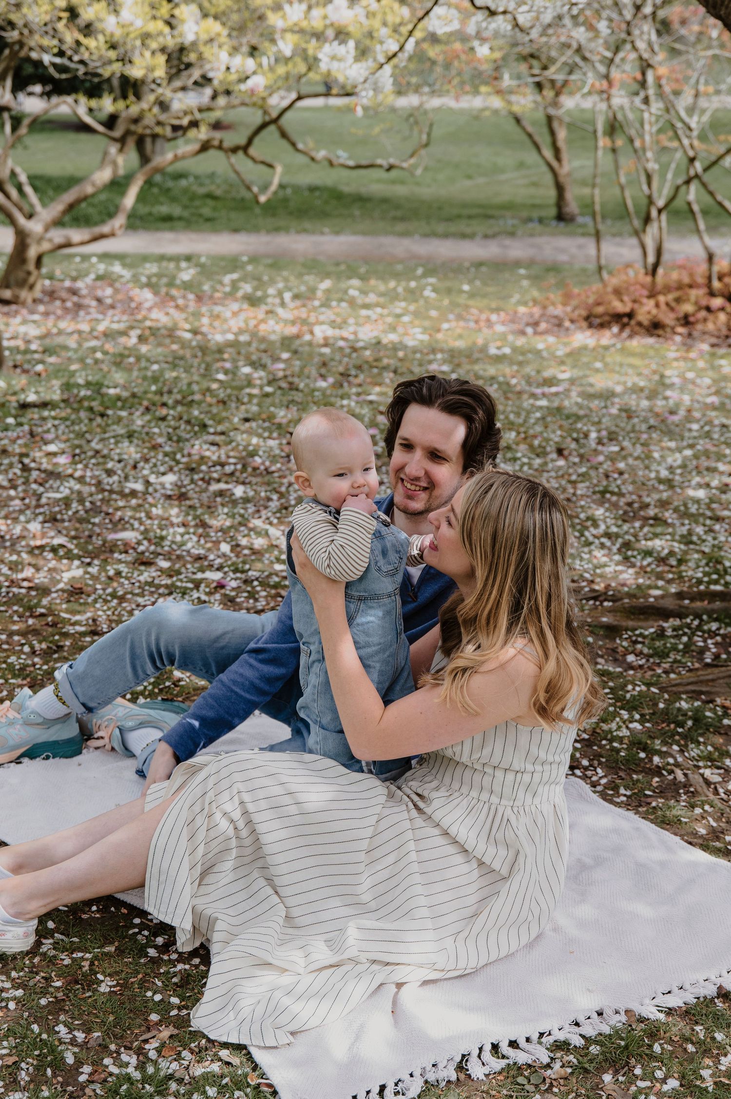 A family enjoys a tender moment together while relaxing on a white blanket spread across fallen leaves in a park setting.