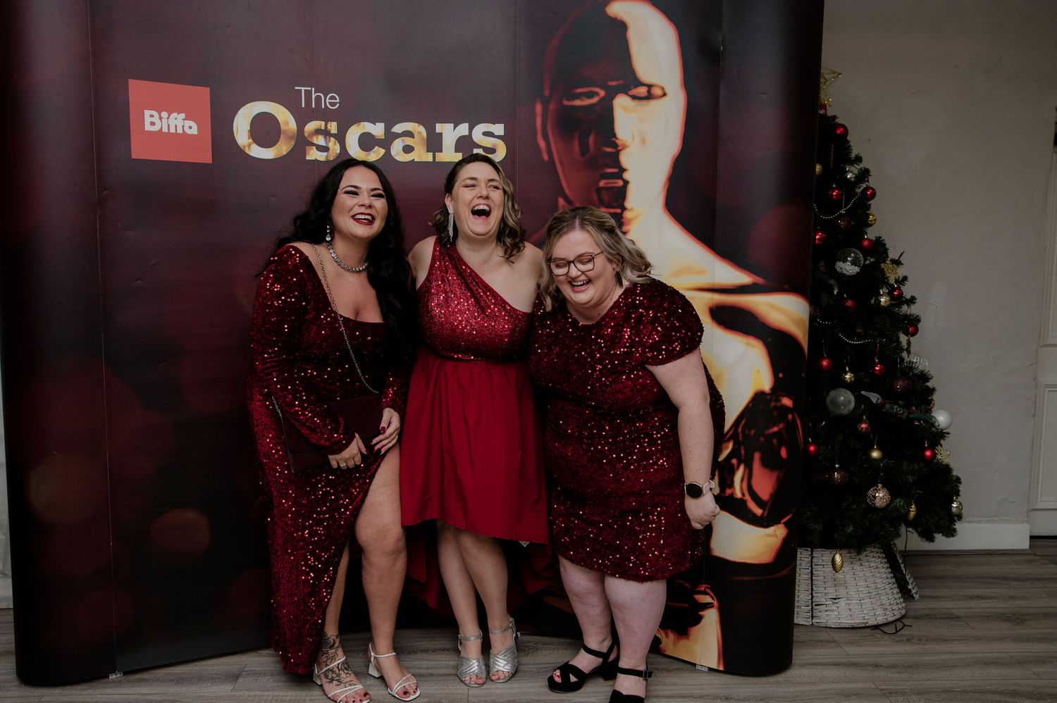 A group in burgundy dresses and black boots pose together at an event with a Christmas tree in the background.