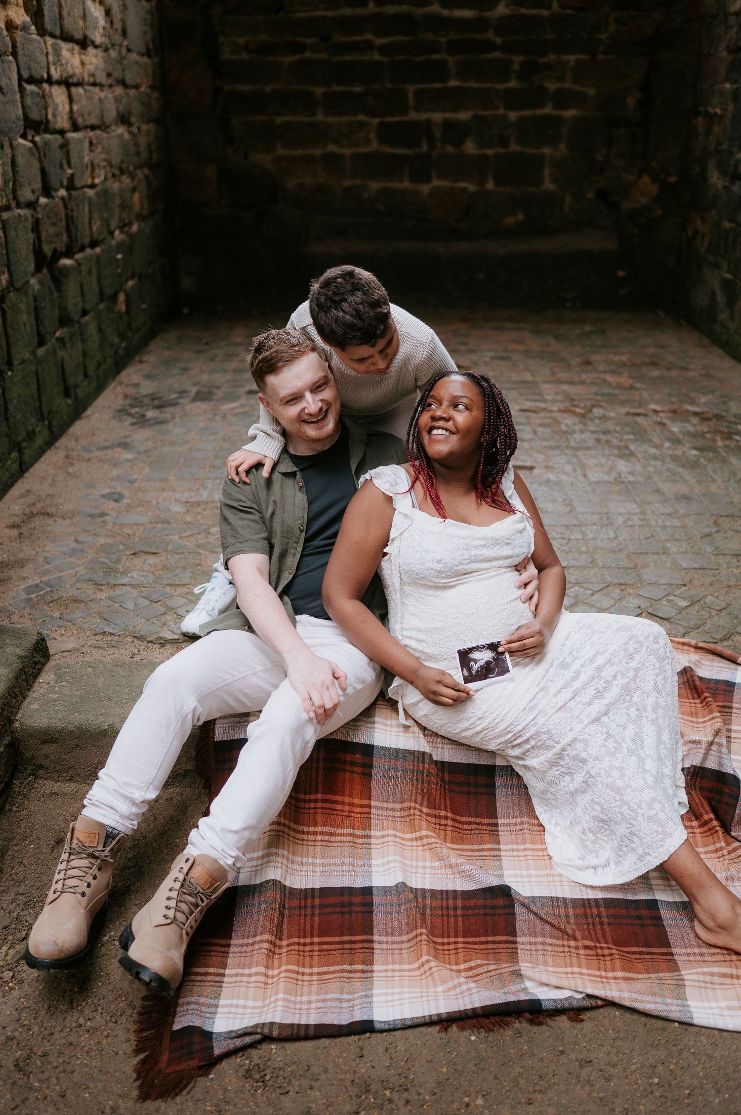 A couple sits on a plaid blanket sharing intimate moments in a brick courtyard setting.
