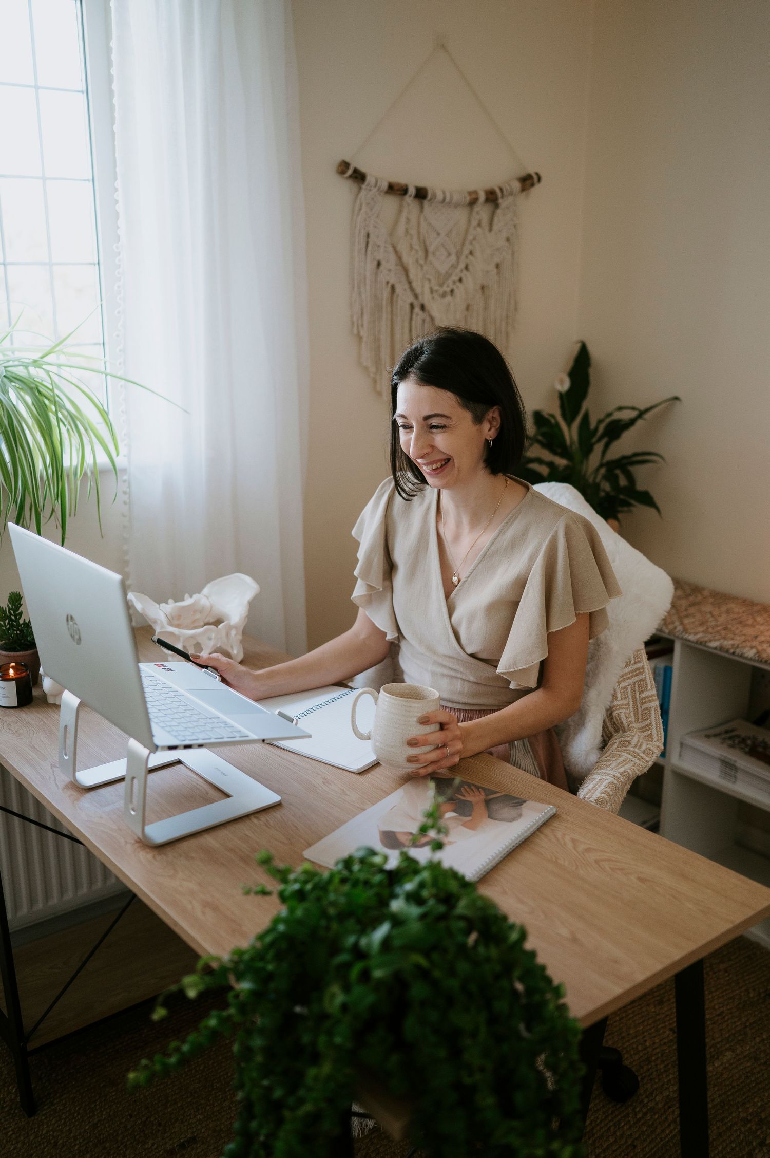 Person in cream colored dress works at wooden desk with laptop and plants in bright minimalist home office setting.