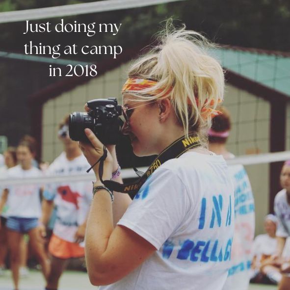 A photographer in a white t-shirt takes photos with a vintage camera at an outdoor event in 2018.