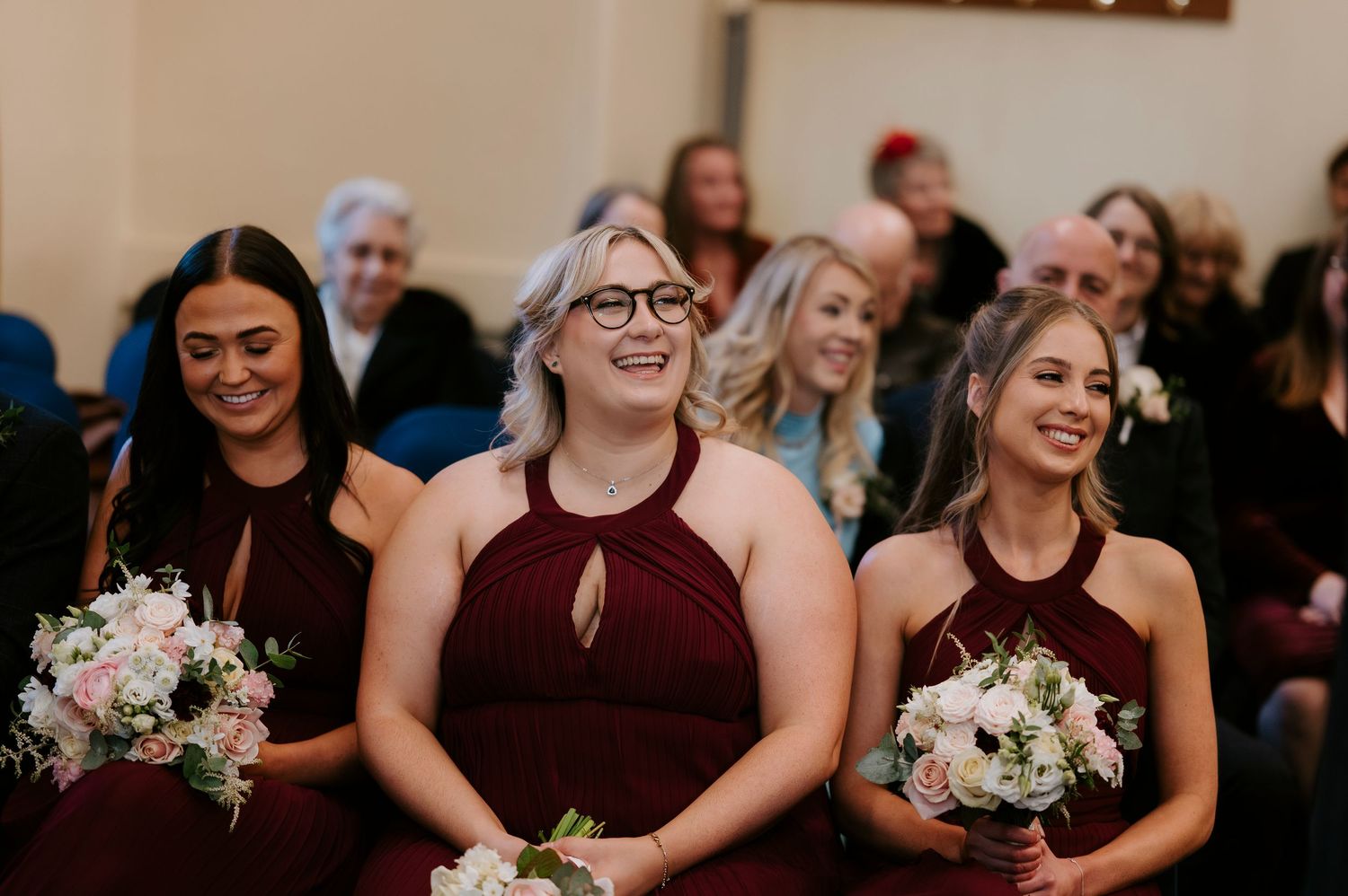 Three bridesmaids in burgundy halter dresses holding bouquets smile joyfully during a wedding ceremony.