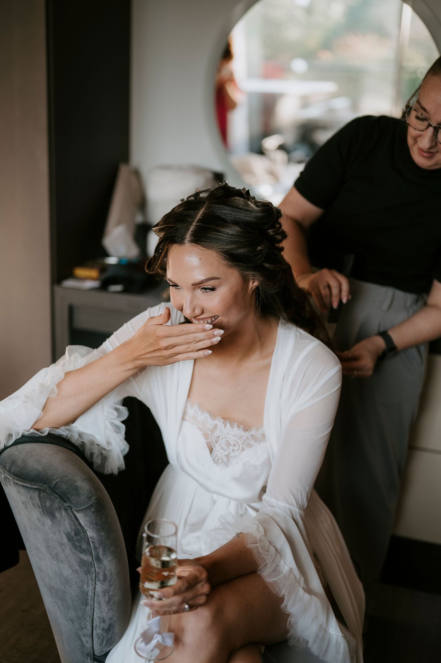 Bride in white robe laughing while holding champagne as hairstylist works on her updo before wedding.