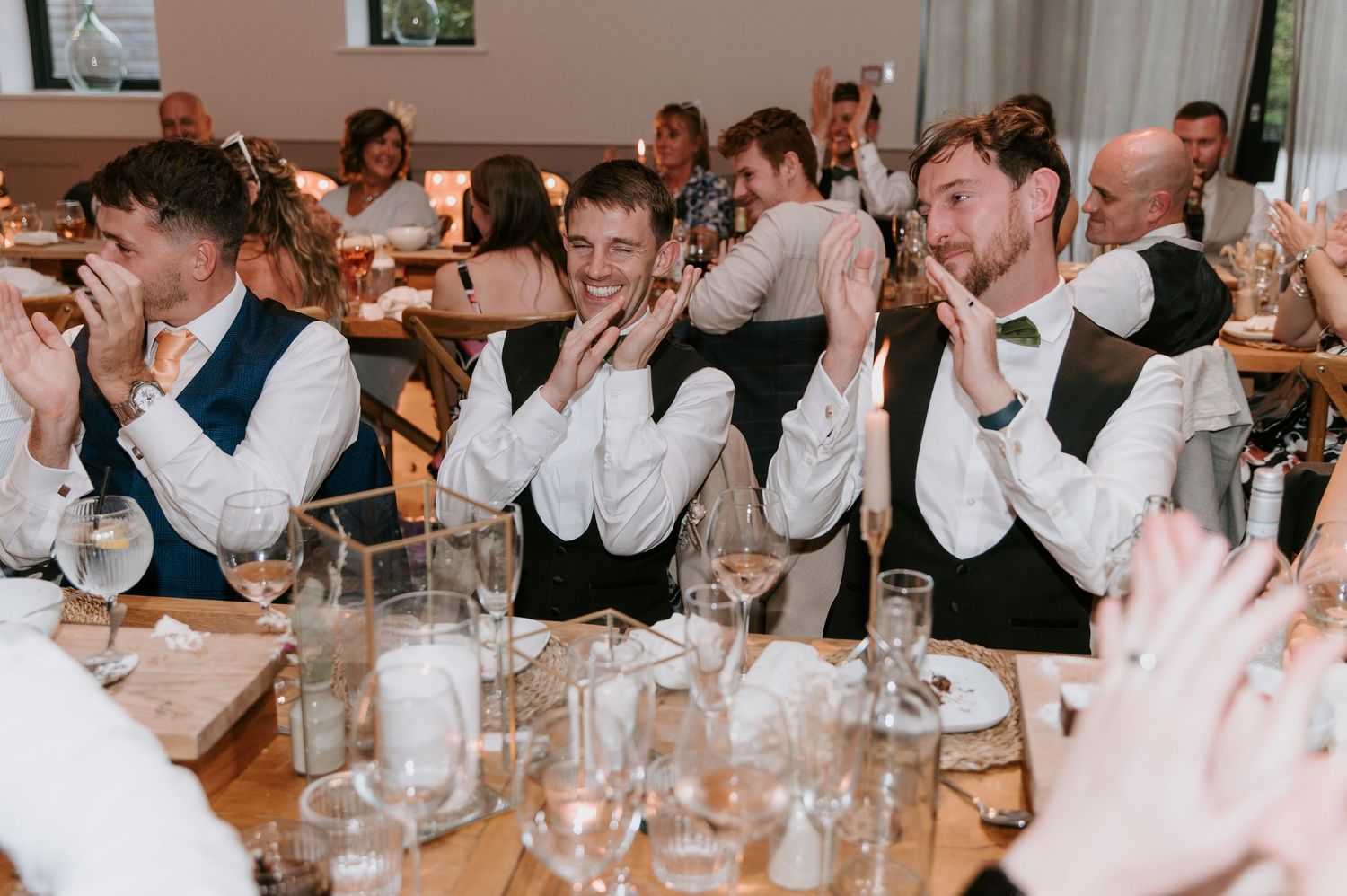 Wedding guests raise their glasses in a toast during a reception dinner at a long banquet table.
