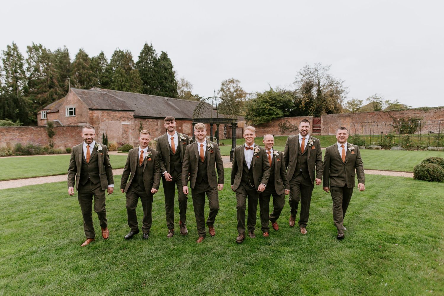 A group wearing dark suits and red ties stands in a line on a grassy lawn next to a rustic brick building.