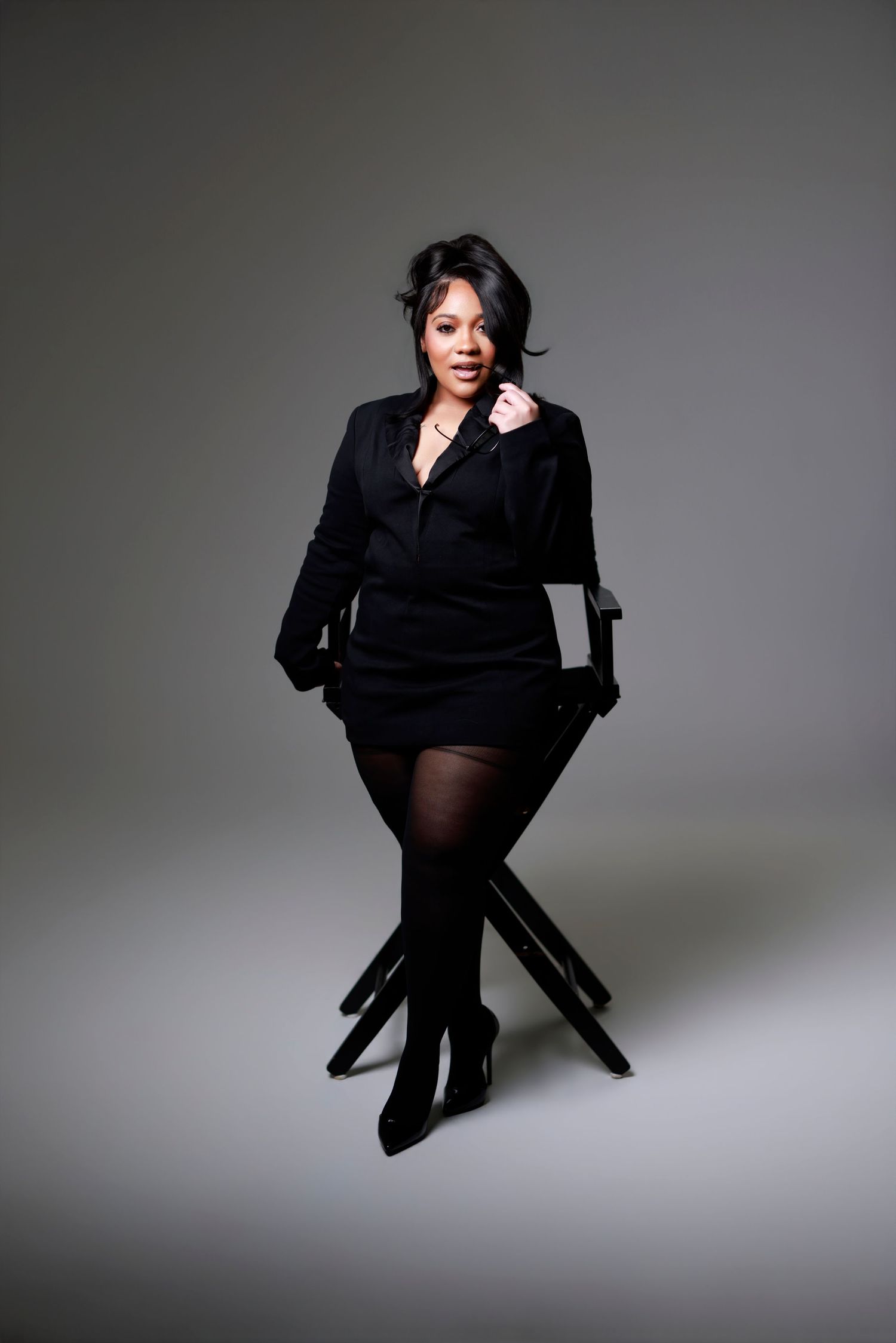Woman in all-black outfit sits on a director's chair against a gray studio background.