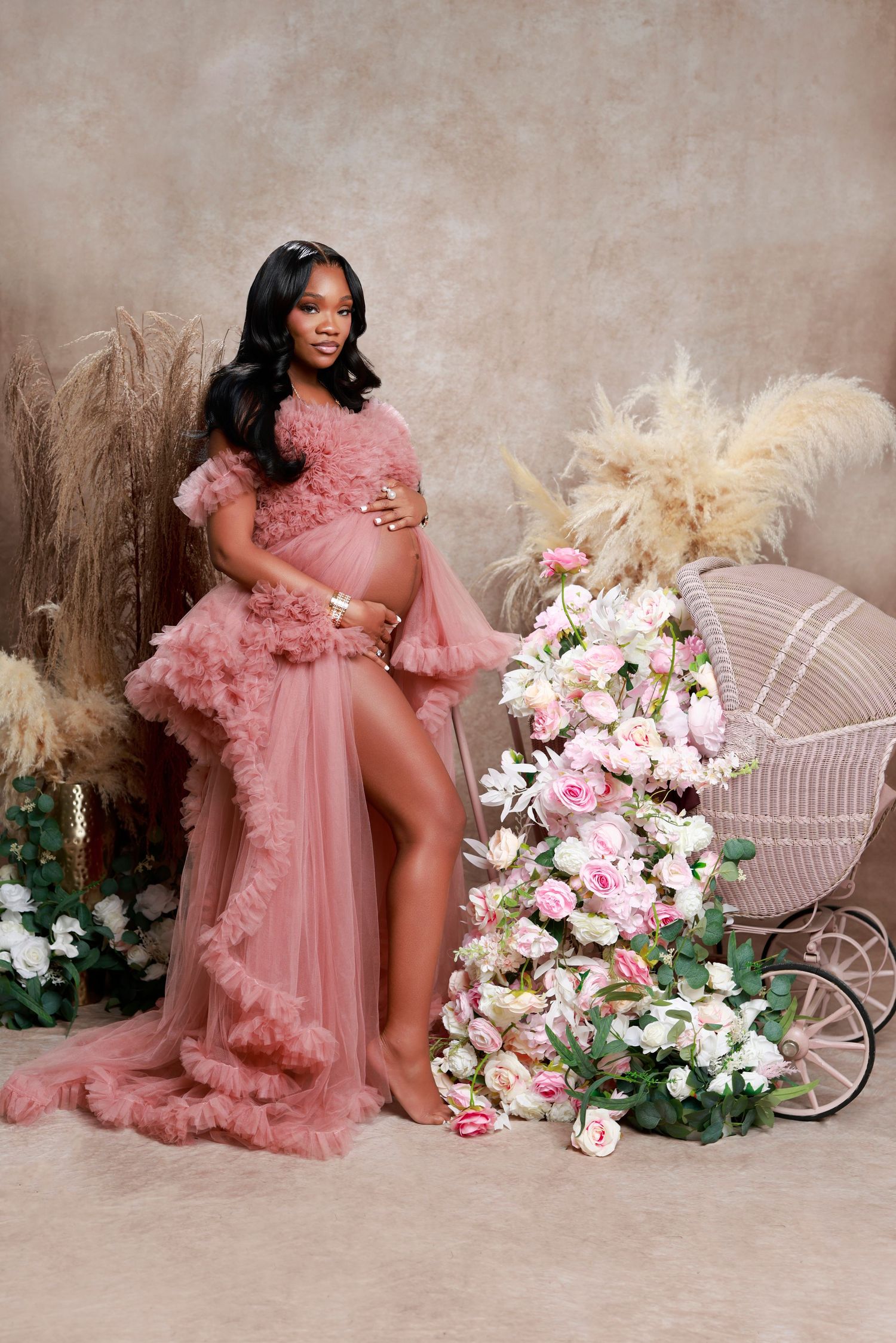 Pregnant woman in pink ruffled gown poses beside floral arrangement and vintage baby carriage.