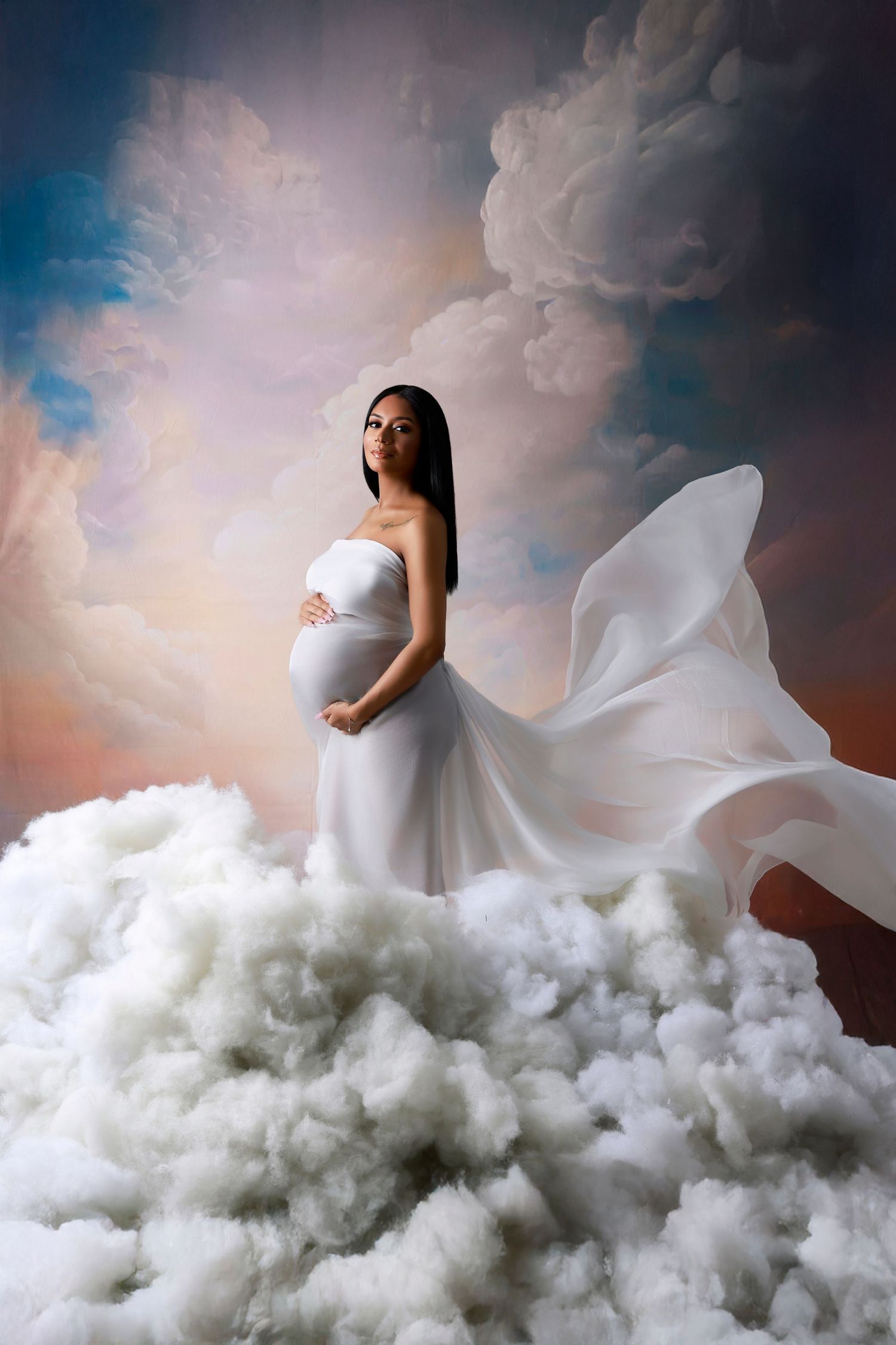 Pregnant woman in white strapless gown stands dramatically among billowing clouds with sky backdrop.